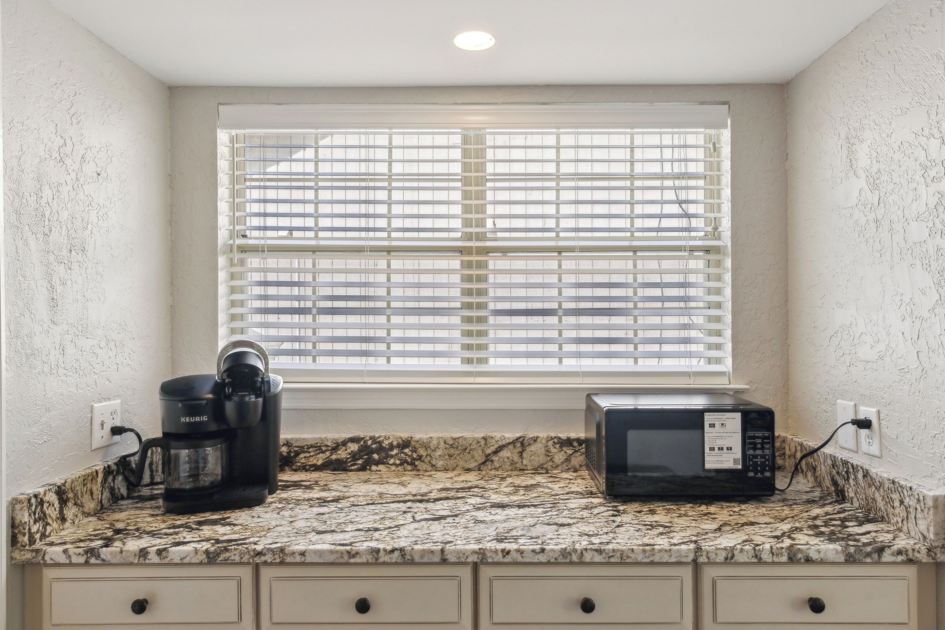Marble patterns swirl across this sleek coffee station, where a modern brewer and microwave wait for your morning routine. Daylight filters through the blinds, highlighting the textured walls and clean, neutral cabinetry.