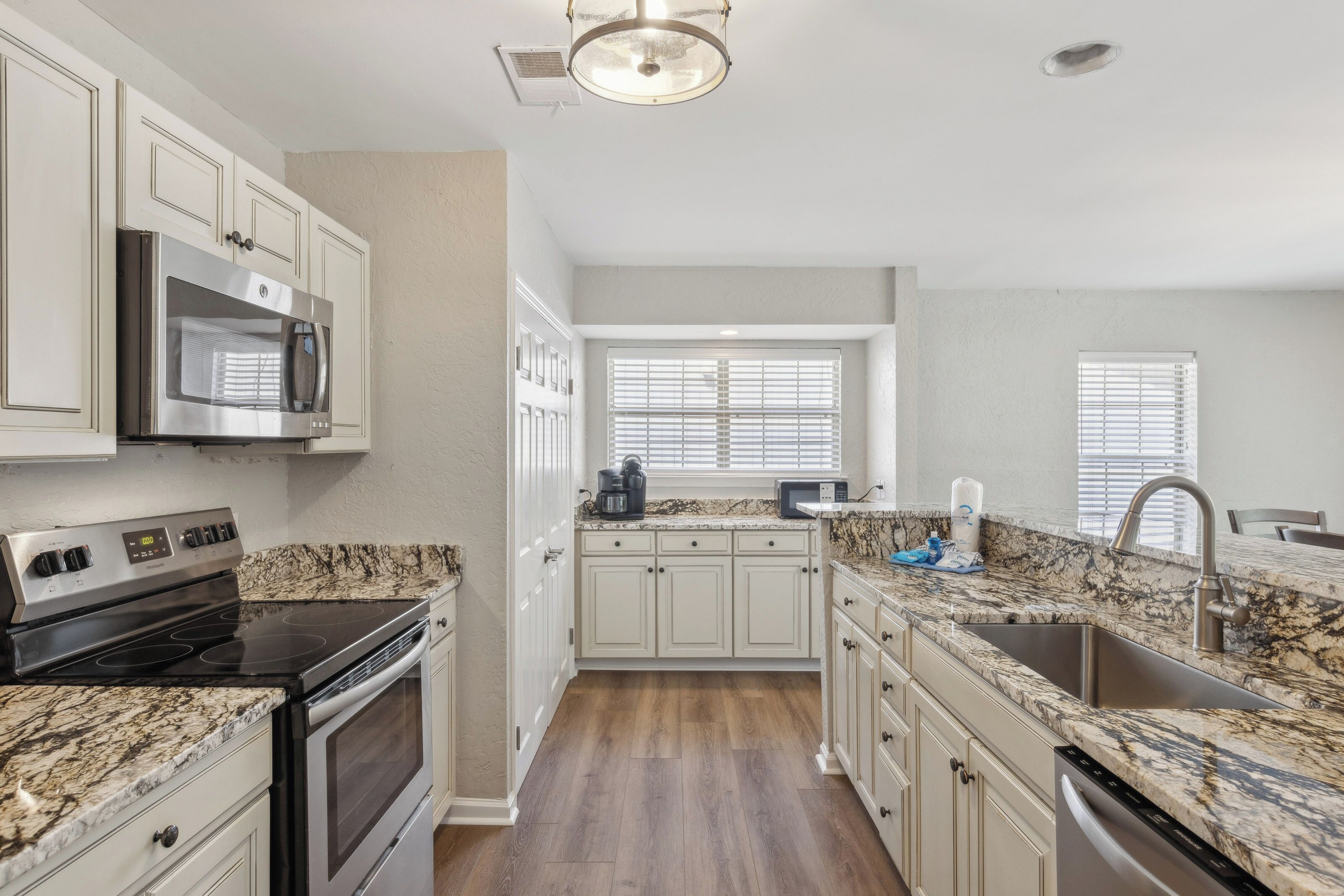 Swirling granite patterns anchor this culinary space, where stainless appliances and crisp white cabinetry create a sleek, efficient layout. Morning rays filter through the blinds to illuminate the wide plank floors and open breakfast bar.