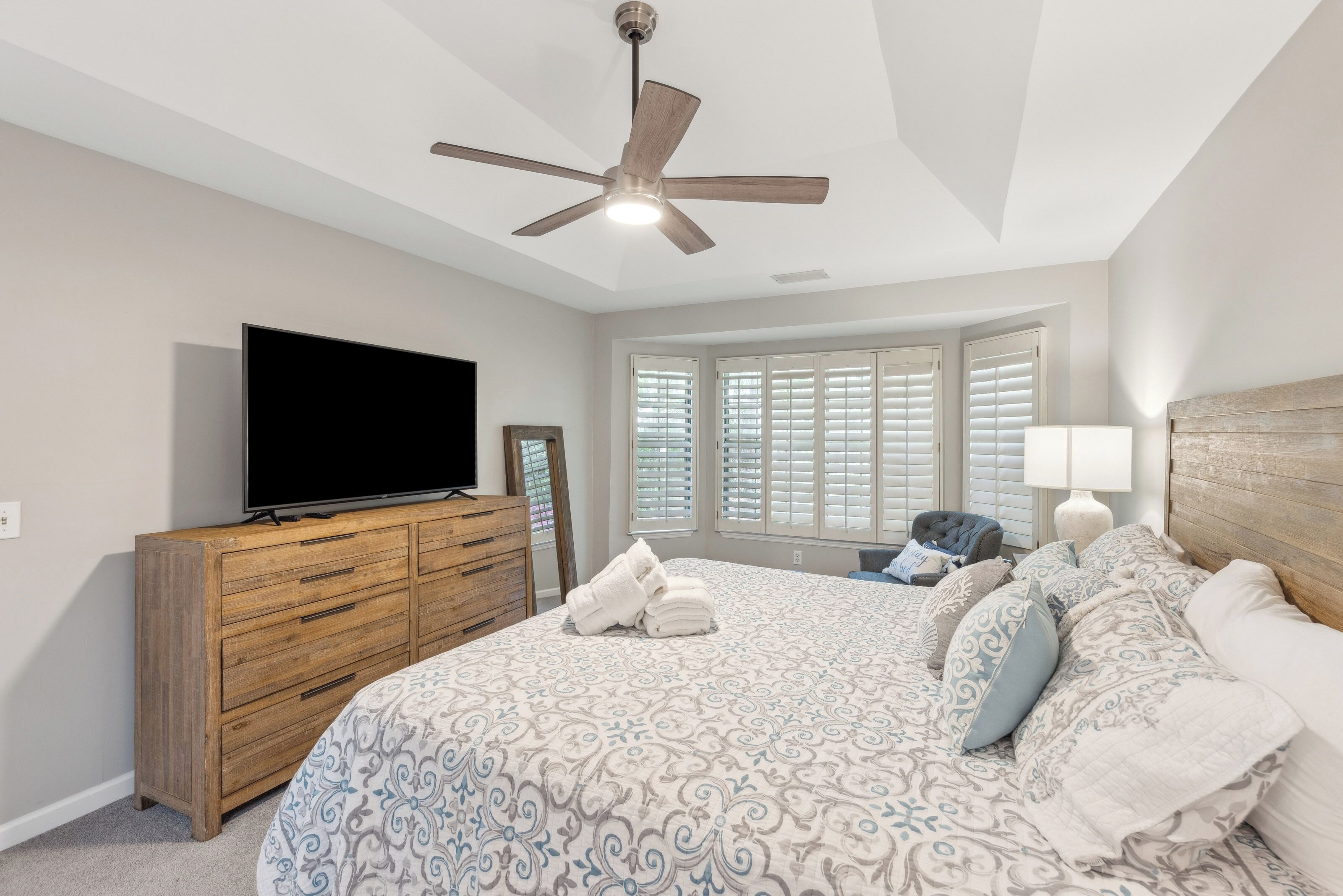 Soft morning glows filter through plantation shutters, illuminating crisp linens and a rustic wood headboard. This quiet bedroom offers a peaceful pocket for rest, defined by a spacious tray ceiling and soothing, neutral tones.