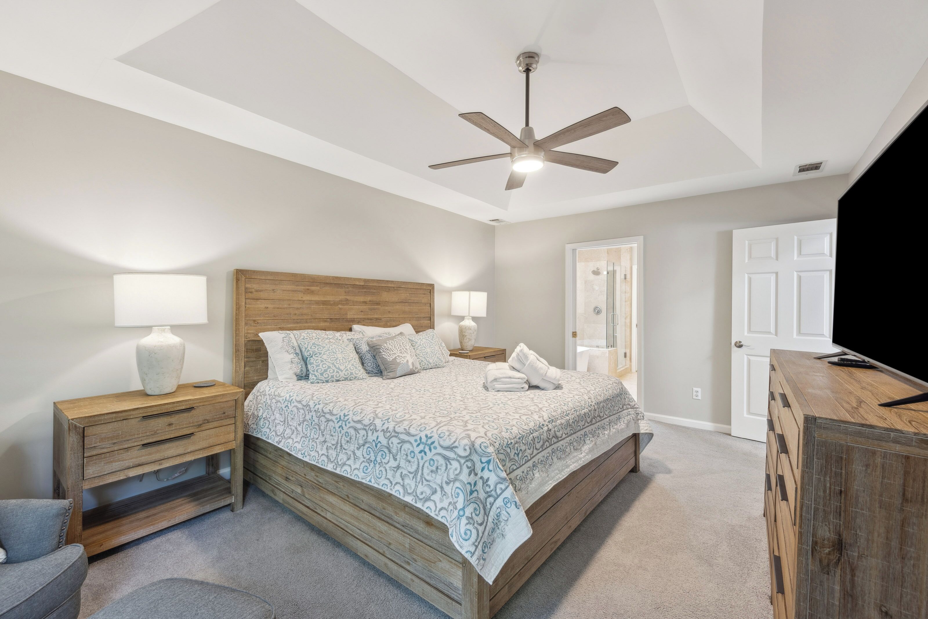 Weathered wood textures and cool, neutral tones create a serene sanctuary for rest. Glowing lamps illuminate the spacious king bed, while a tray ceiling and overhead fan keep the room feeling light and open for a peaceful night.