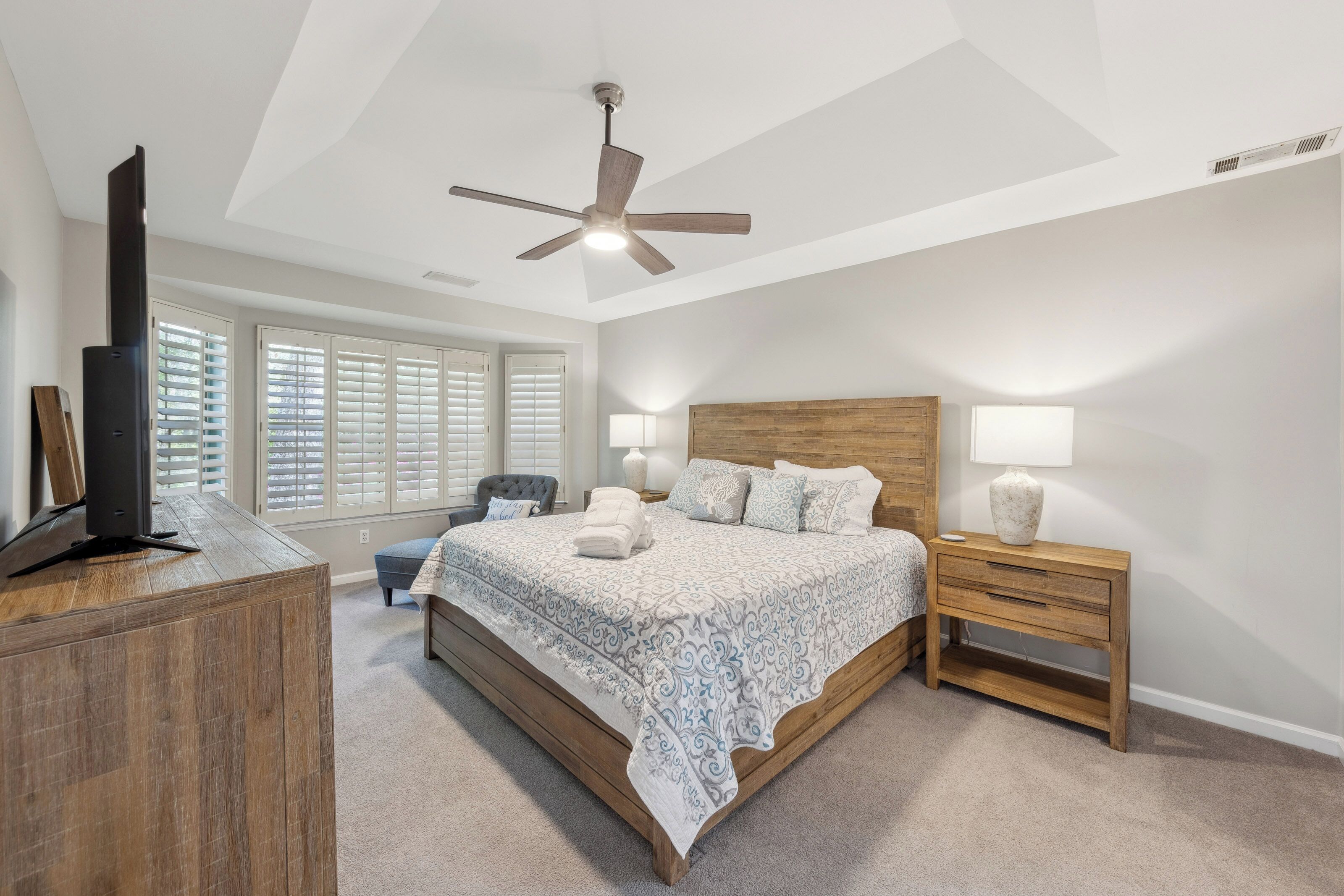 Reclaimed wood textures and soft linens create a serene sanctuary beneath a recessed tray ceiling. Gentle afternoon glow filters through plantation shutters, highlighting the cozy velvet armchair and spacious, cloud-like bed.