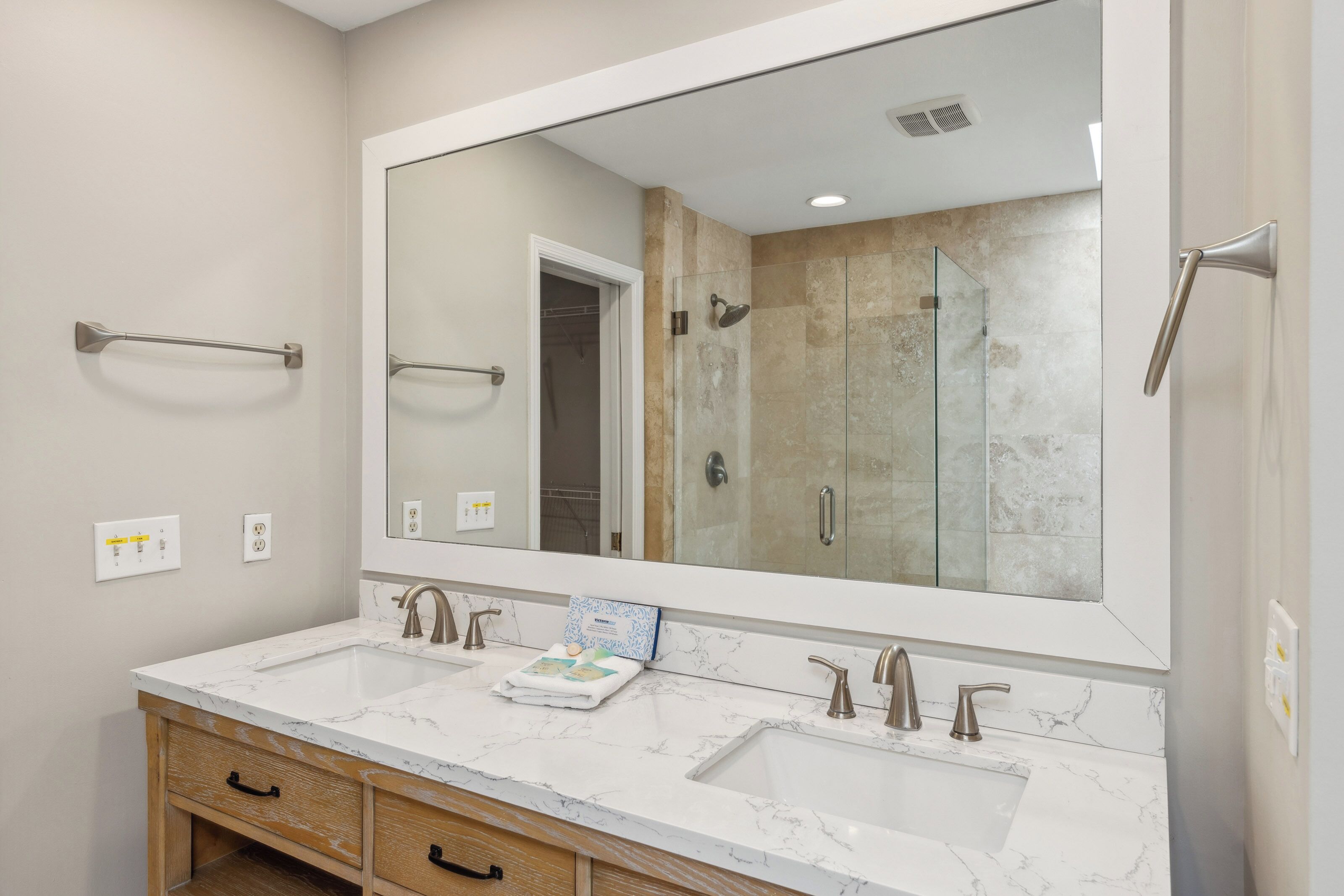 Honey-toned wood cabinetry grounds this spa-like vanity, topped with a crisp white quartz counter and dual basins. A glass-walled shower reflects in the oversized mirror, creating a sense of quiet luxury for your morning routine.