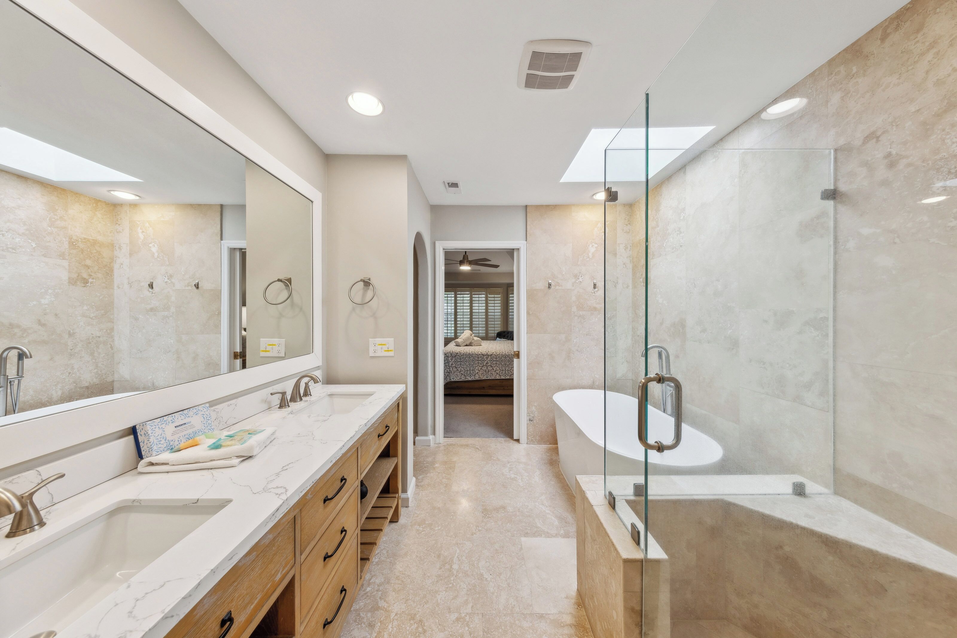 Morning light pours through the skylight, illuminating the crisp white soaking tub and textured tile. A dual vanity with oak accents offers ample room for your morning routine in a space designed for calm and clarity.