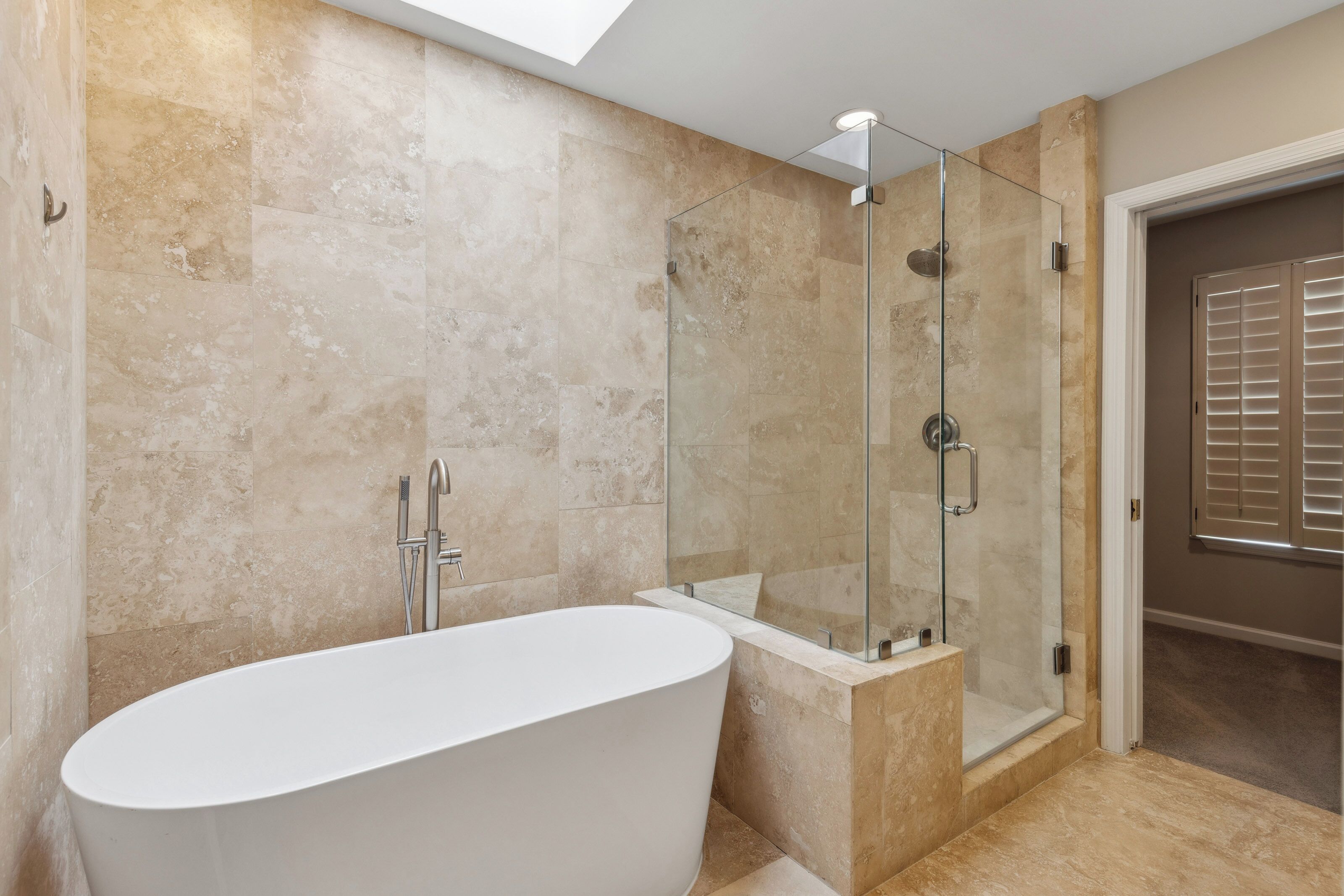 Travertine tiles wrap this spa-like sanctuary in organic texture, complemented by a deep soaking tub and a sleek glass shower. A hidden skylight pours a midday glow over the clean lines and brushed nickel finishes for a peaceful morning routine.