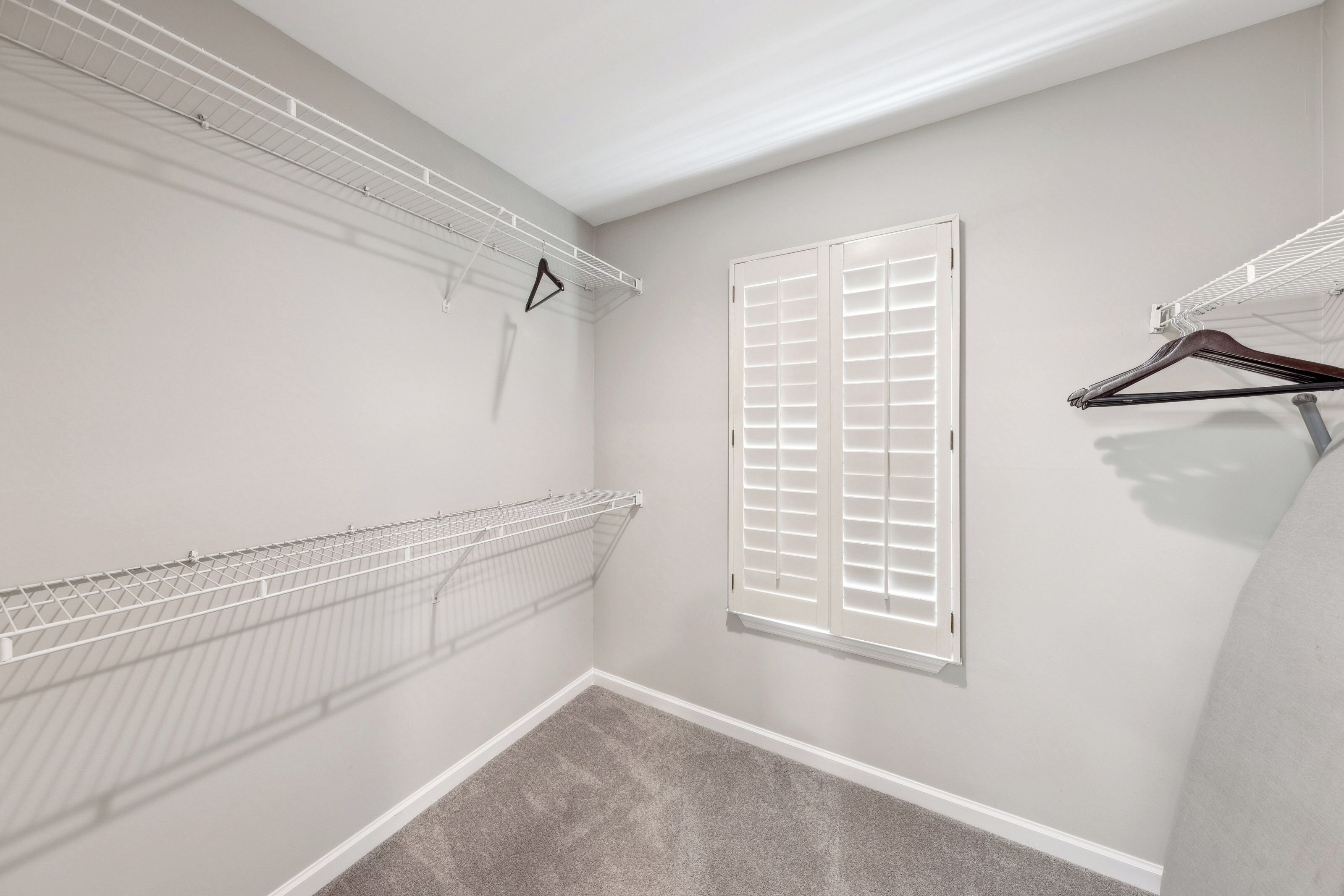 White plantation shutters filter a gentle glow into this spacious walk-in closet. Crisp wire shelving and plush gray carpeting create an organized, calm environment for unpacking your favorite travel outfits.