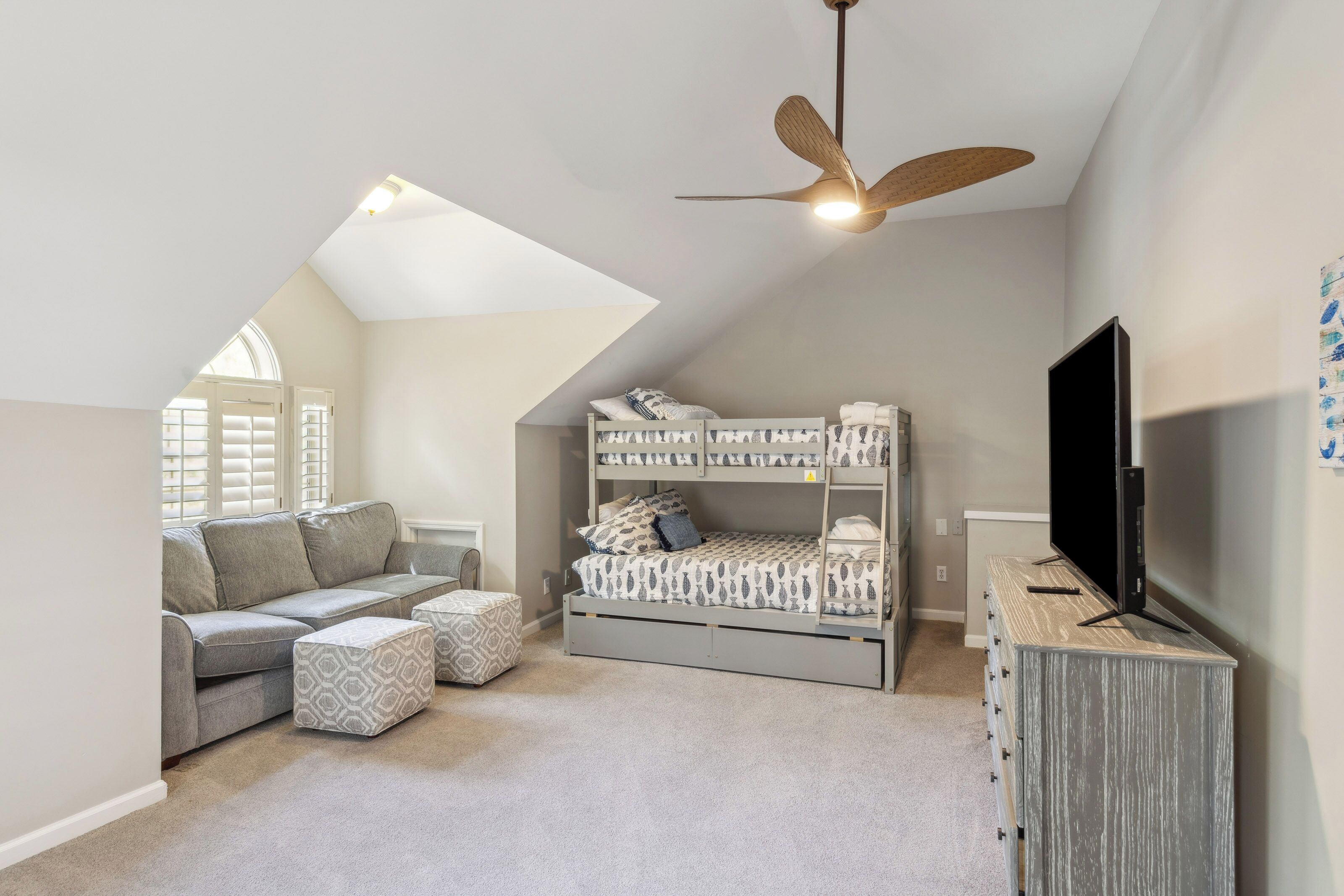 Sloping ceilings and a wood-bladed fan create a cozy, tucked-away feel in this playful loft. Modern bunk beds and a deep sofa offer a comfortable spot for movie nights or quiet afternoons under the gentle glow of the arched window.
