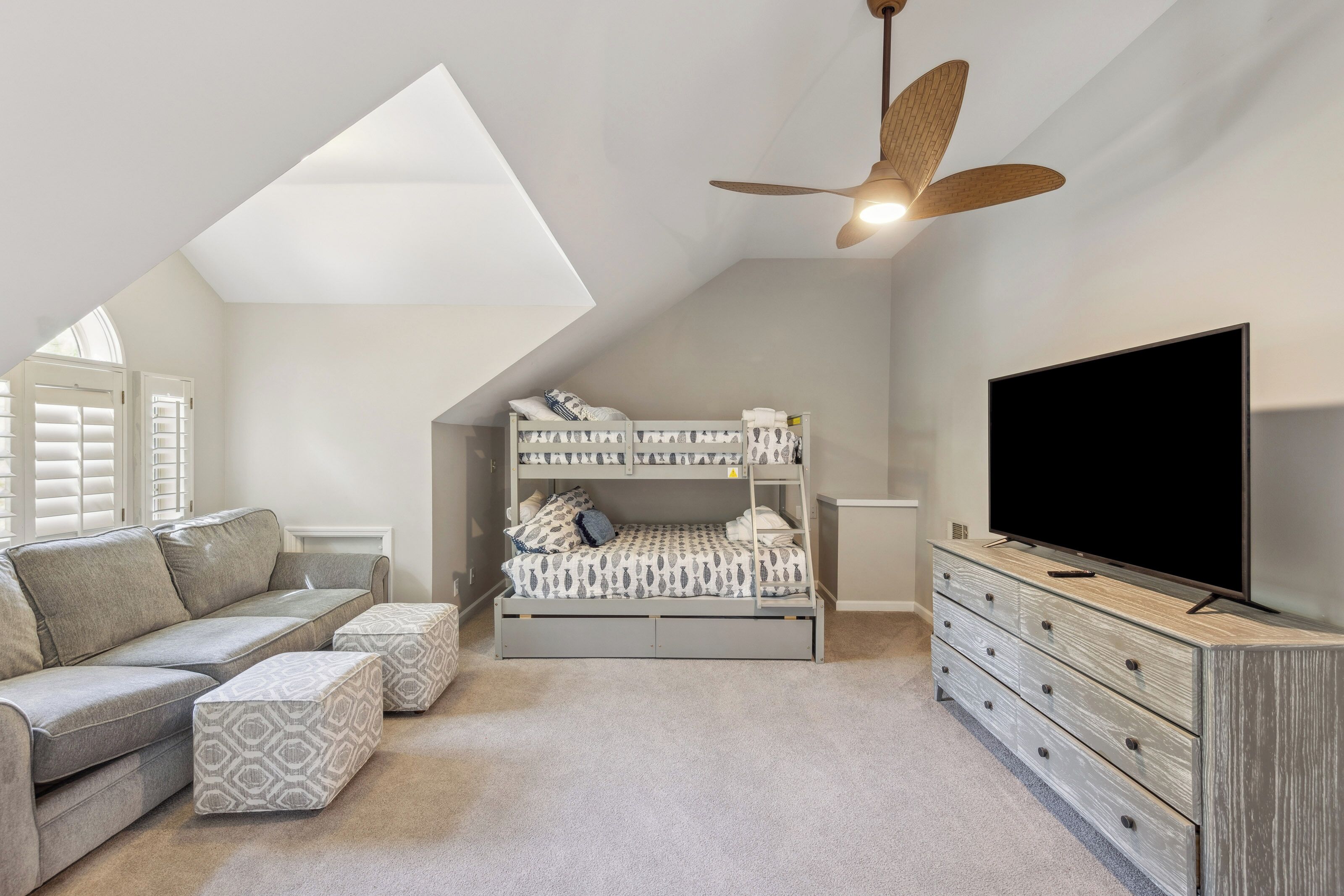 Textured grey fabrics and wood tones create a cozy hideaway under the sloped ceilings. Day light spills through the shutters, illuminating a playful bunk bed station and a spacious media corner perfect for a quiet family movie night.
