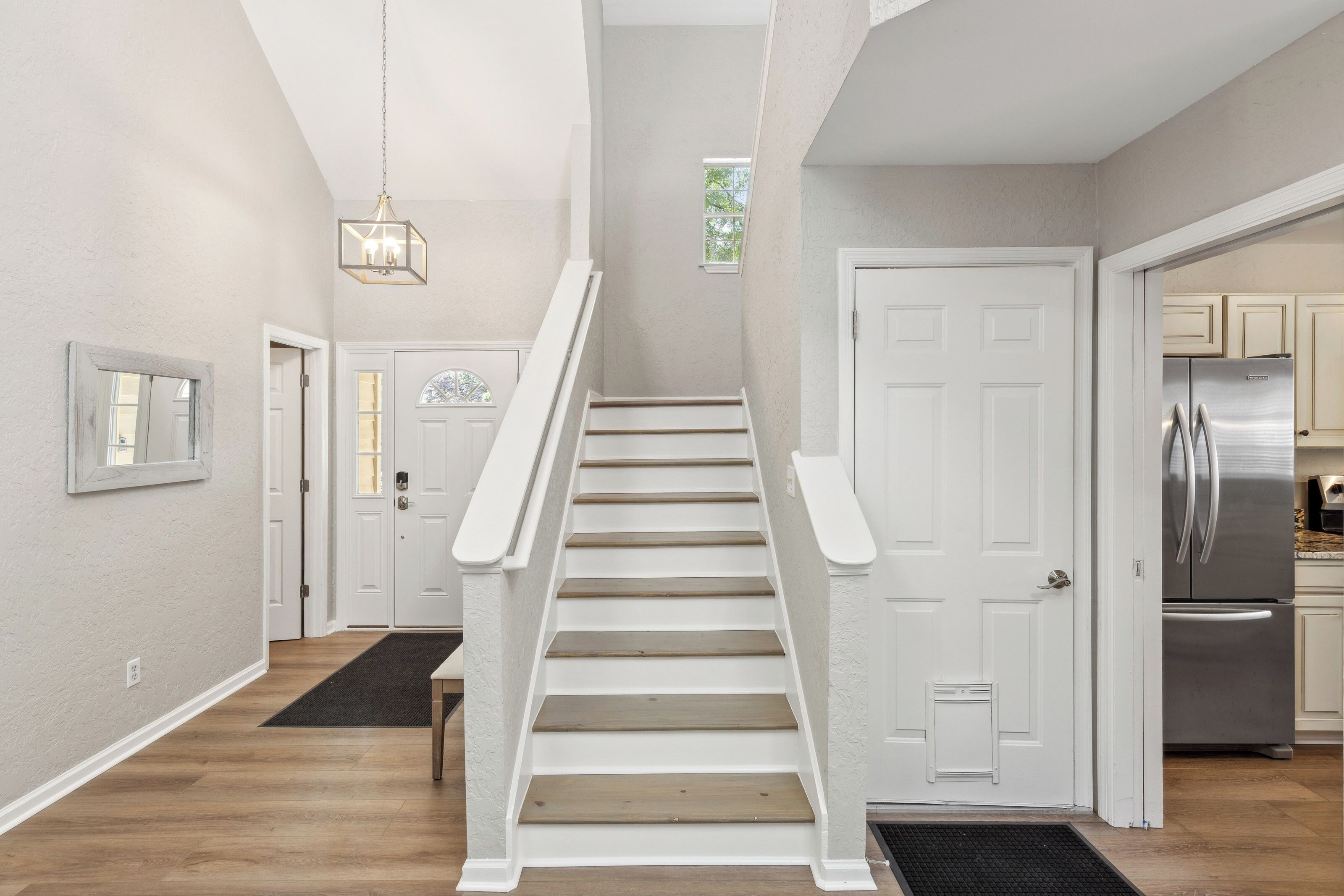 Oak-toned treads and crisp white risers guide the eye upward, framed by a soaring entryway and a modern lantern pendant. High ceilings and a clean, neutral palette create a sense of calm and openness the moment you arrive.