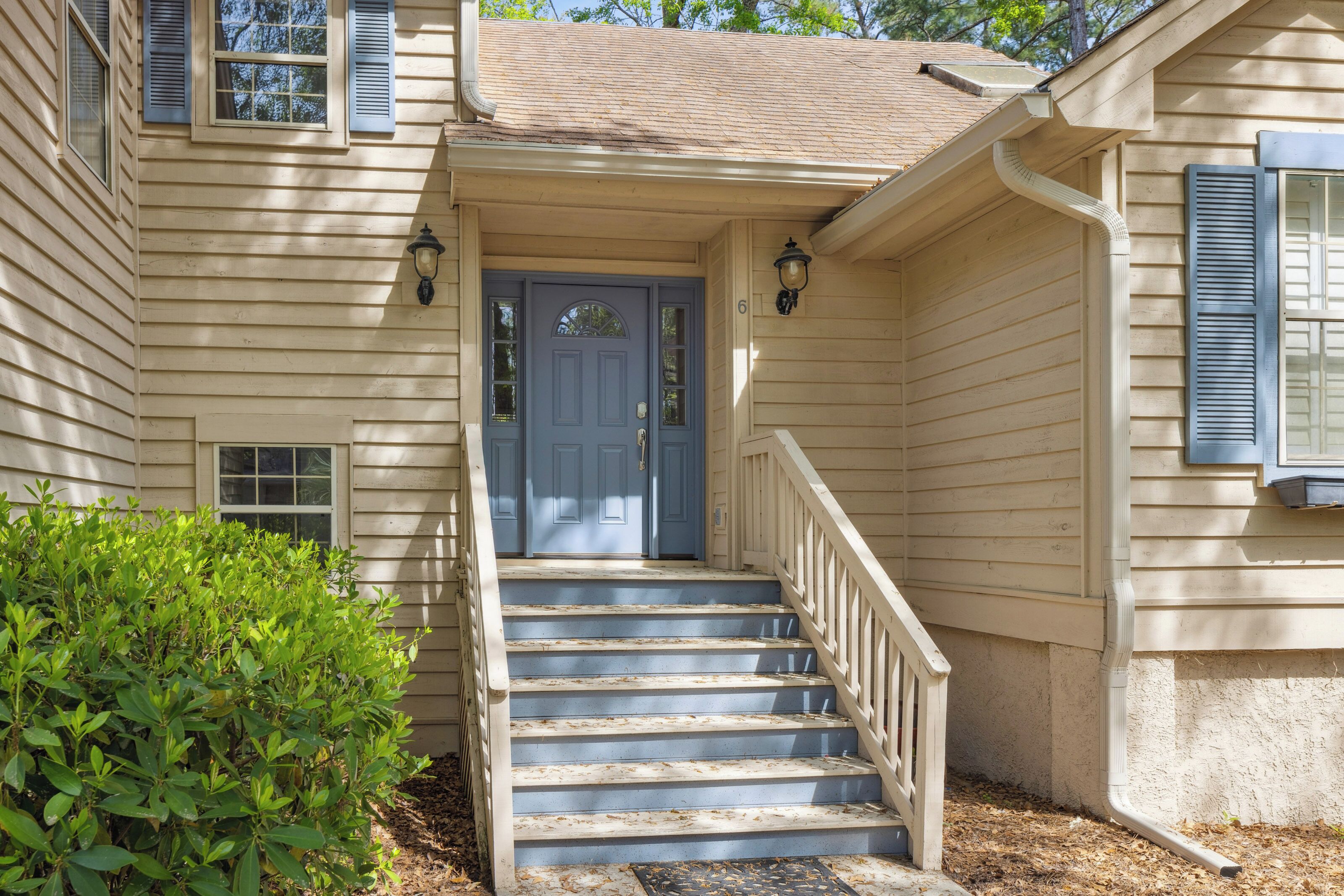 Blue accents and creamy siding glow in the afternoon radiance, creating a cheerful greeting. These wooden stairs lead you to a quiet home where soft shadows and lush greenery frame your arrival.