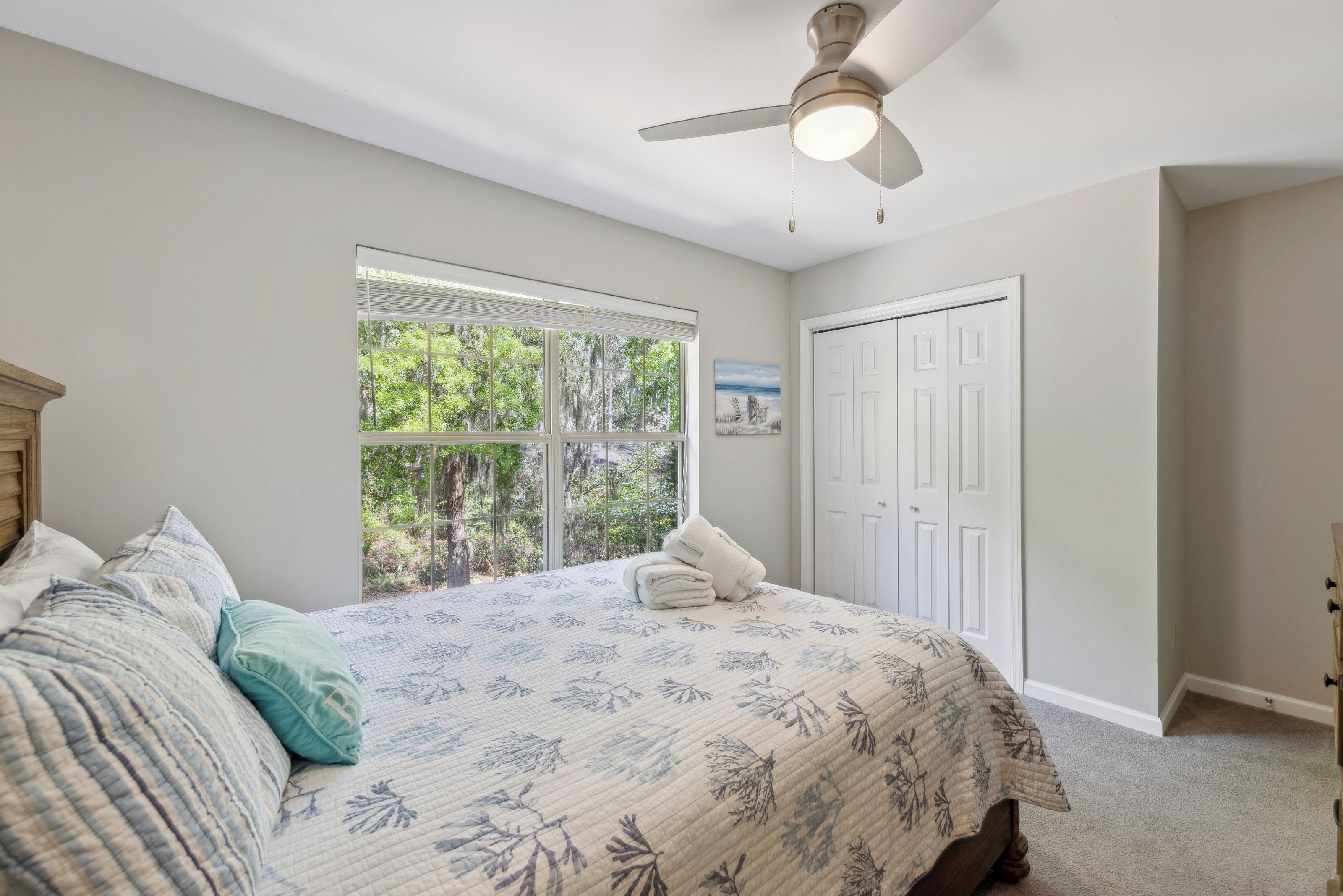 Daylight pours through wide windows to illuminate this serene bedroom, highlighting the coral-patterned linens and cool seafoam accents. Lush green views create a peaceful backdrop for a restful afternoon nap.