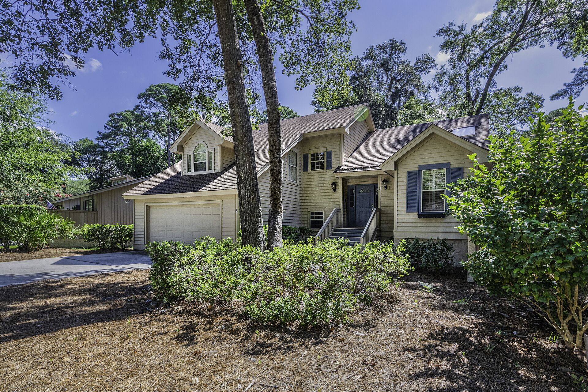 Tall oaks cast dancing shadows across this quiet cedar-shingled home, where dappled morning rays filter through the pine needles. A slate-blue door and matching shutters offer a calm, coastal greeting beneath the bright Carolina sky.