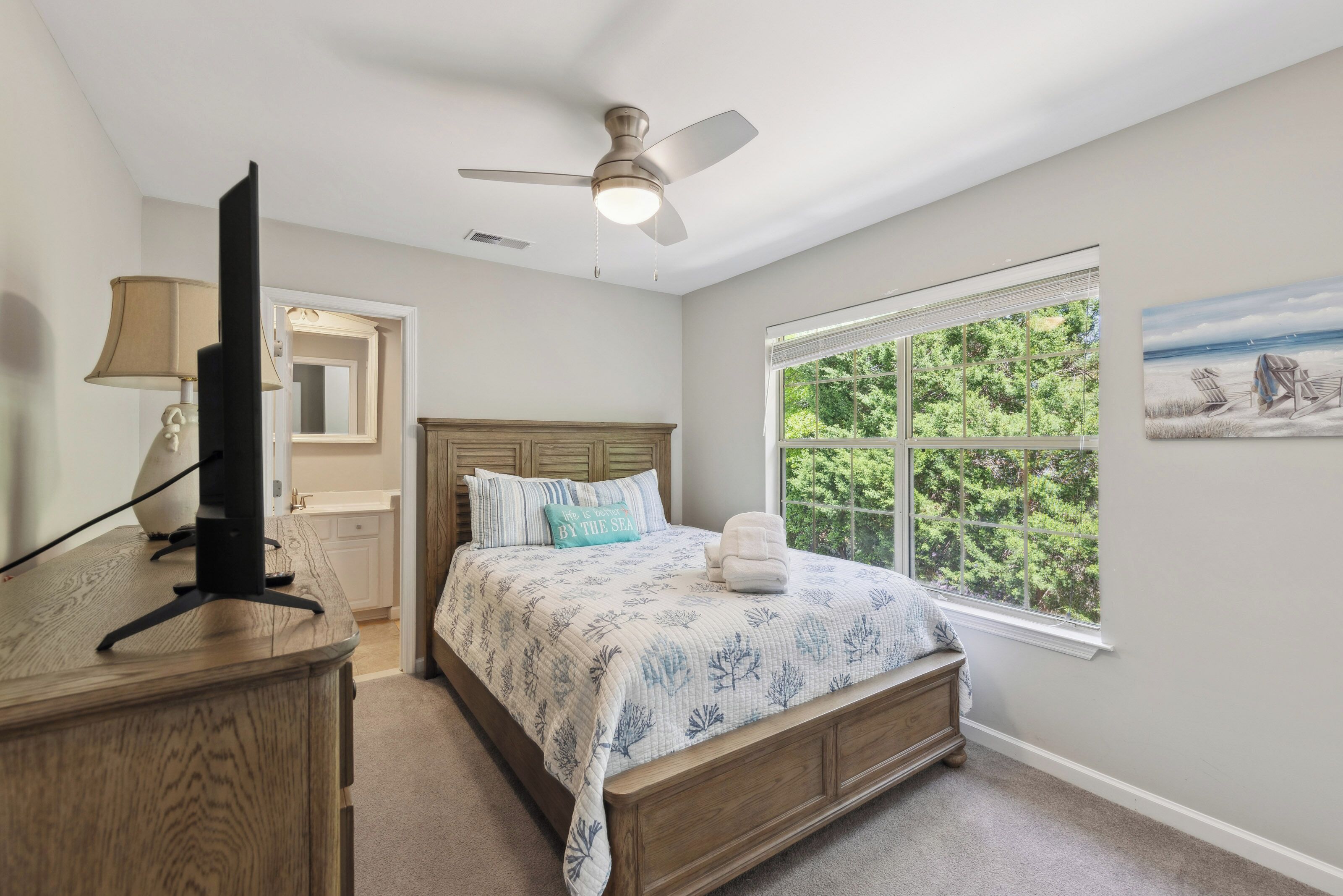 Subtle coastal tones and wood grain finishes create a serene sleep space, where garden views spill through the wide window. Leafy greens frame the bedside, while a soft breeze from the ceiling fan keeps the afternoon air perfectly cool.