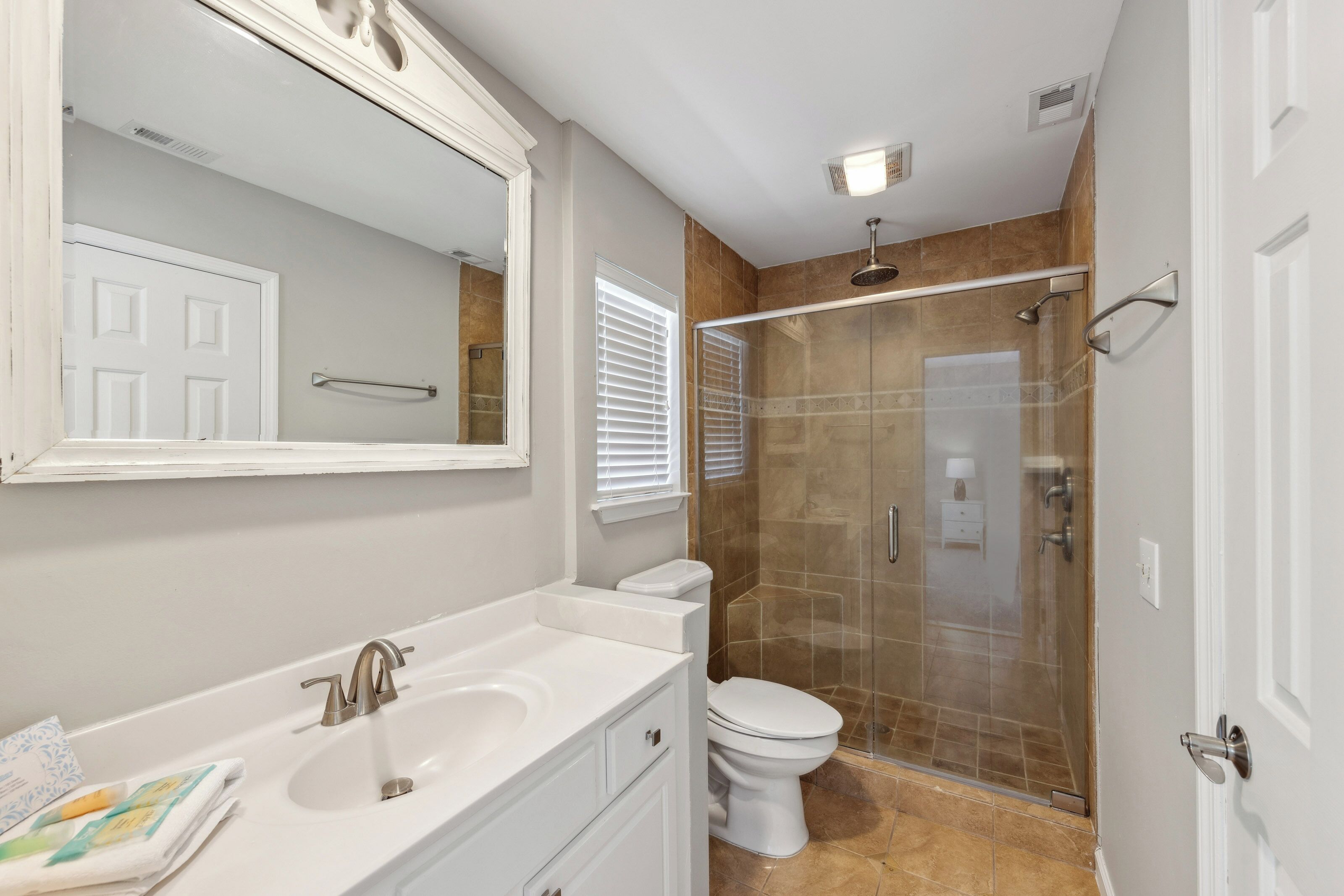 Earthy stone tiles and a spacious glass shower create a spa-like atmosphere for your morning routine. Bright white cabinetry and a vintage-style mirror catch the glow of the overhead light, offering a clean and polished space to refresh.