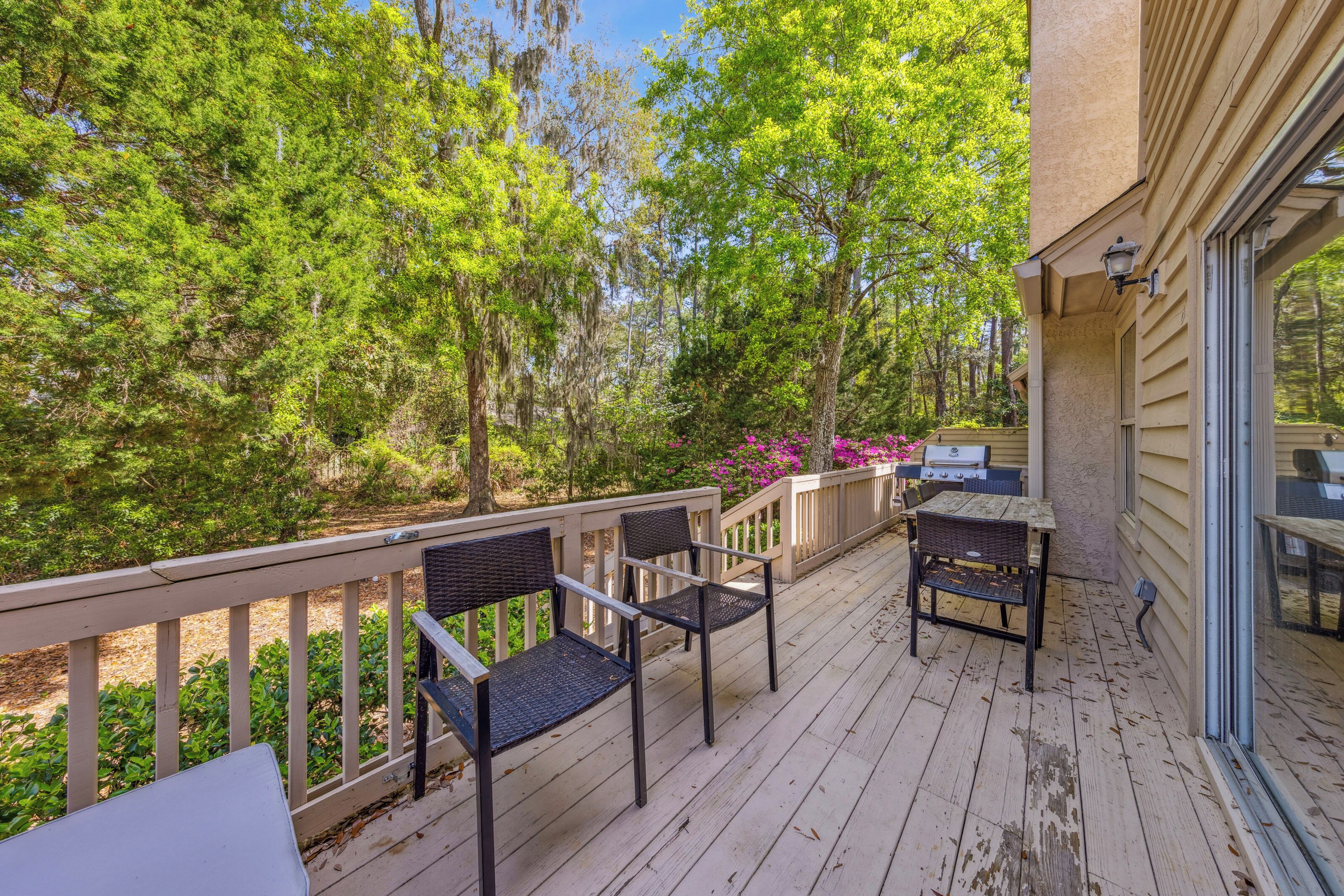 Weathered wood planks lead the way to a peaceful outdoor dining setup framed by lush, towering trees and vibrant pink blooms. This secluded deck offers a quiet spot for morning coffee or evening grilling in the dappled afternoon glow.