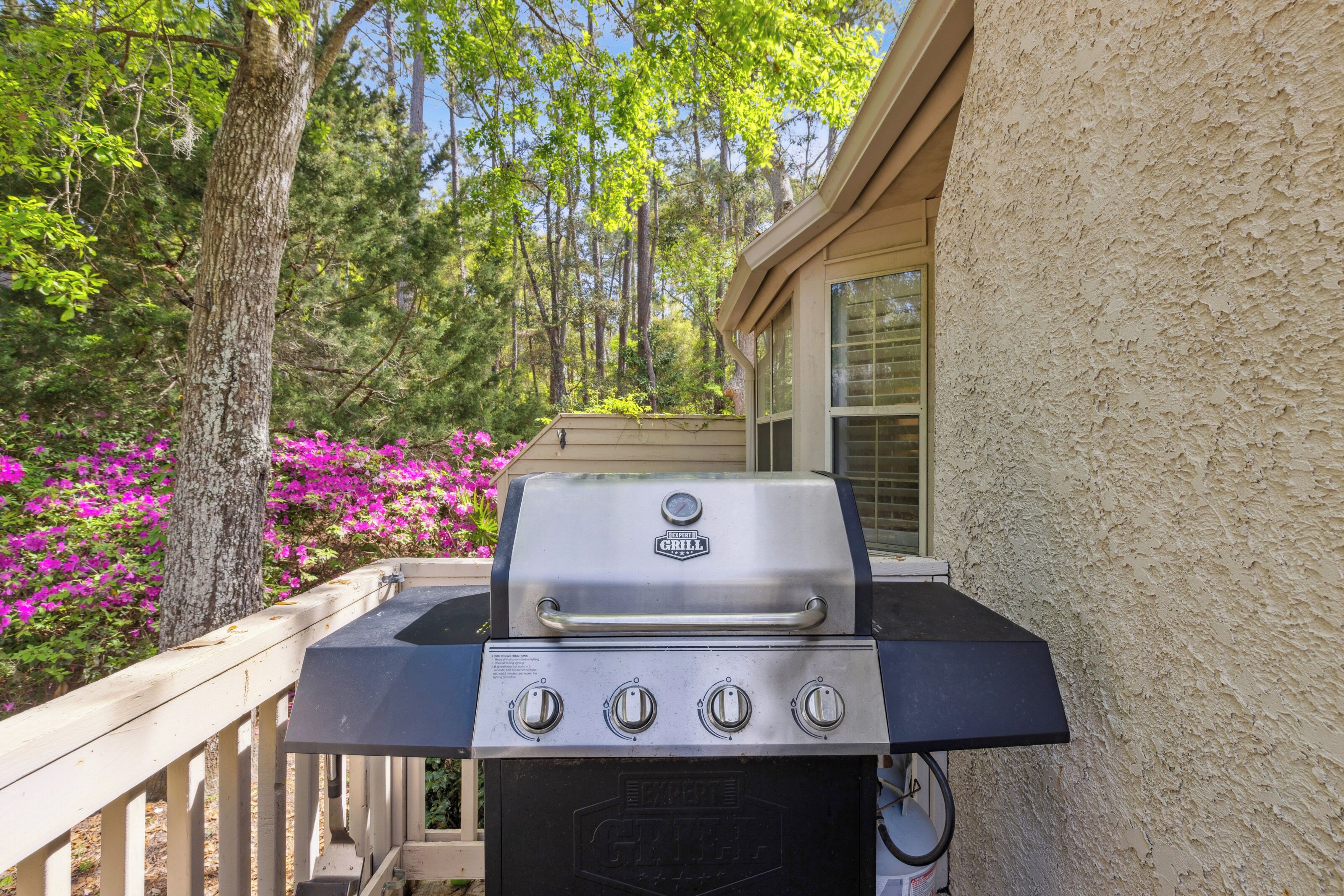 Vibrant azaleas frame this private deck, creating a colorful backdrop for afternoon cookouts. The sleek grill sits ready for an alfresco dinner under a canopy of towering trees and clear blue skies.