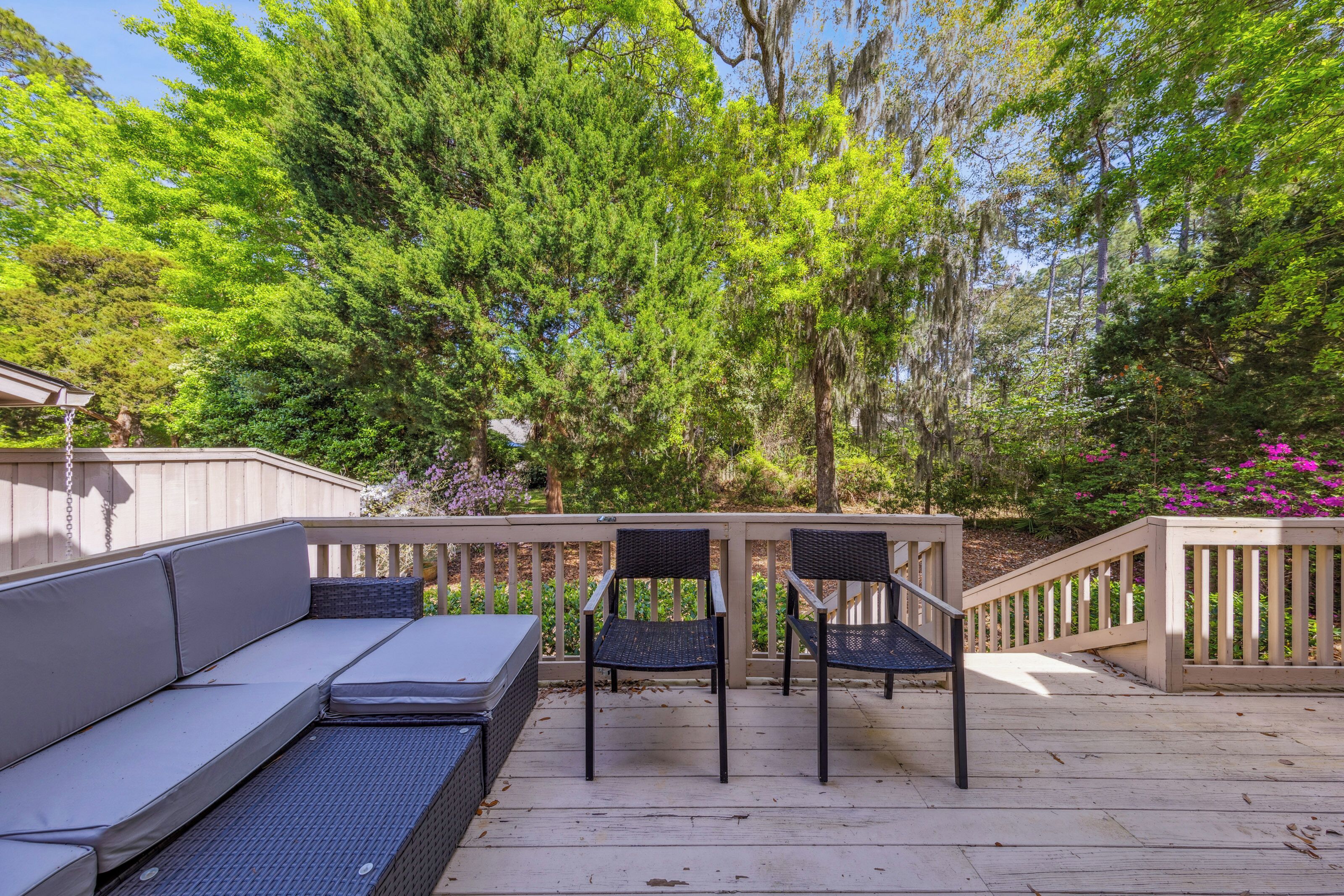 Woven wicker chairs and plush cushions sit ready on this sun-drenched wooden deck. Towering green trees and hanging moss create a vibrant, private backdrop for your morning coffee or a quiet afternoon spent in the fresh air.