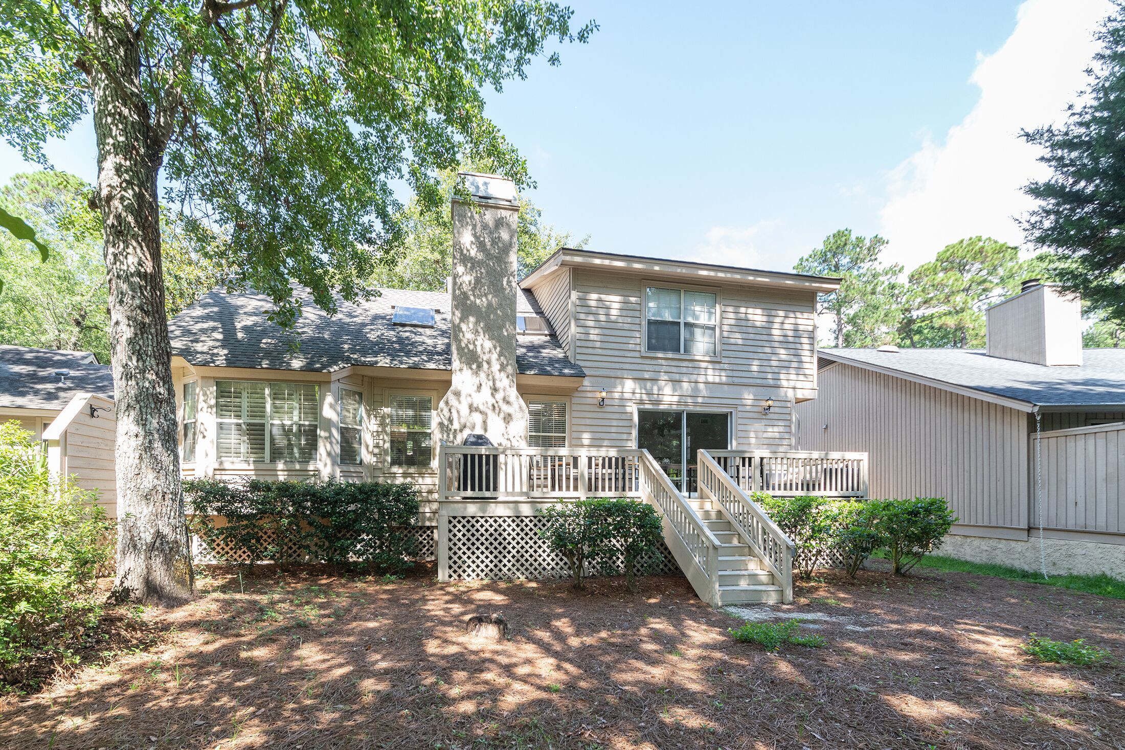 Leafy canopies cast dancing shadows across this quiet backyard, where a spacious wooden deck invites slow afternoons in the fresh air. The neutral siding and stone chimney blend into the forest, creating a calm home for your next getaway.