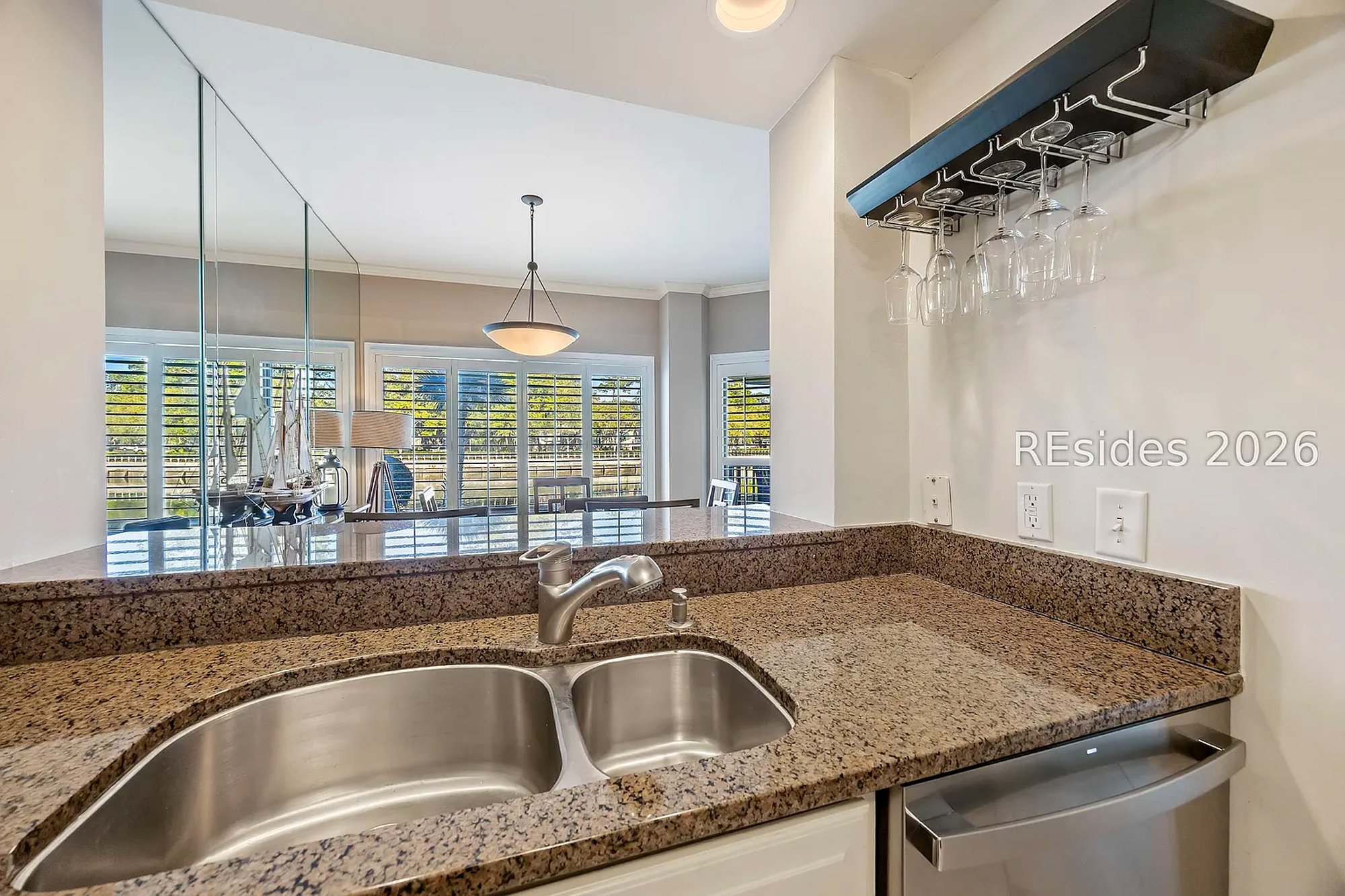 Stone counters shimmer under the soft glow of the hanging bar glassware, creating an ideal spot for evening drinks. Through the open kitchen pass, afternoon beams filter through white shutters to illuminate the airy dining space.