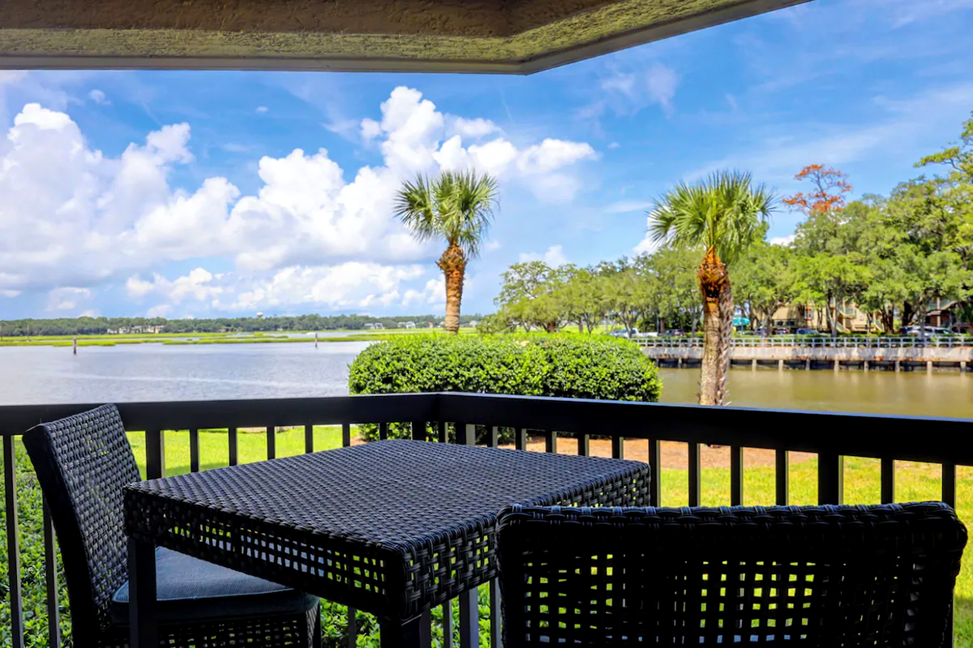 Woven outdoor seating faces a bright waterfront horizon, where palms sway under a vast blue sky. Sip coffee in the afternoon glow while watching the tide roll past this quiet perch.