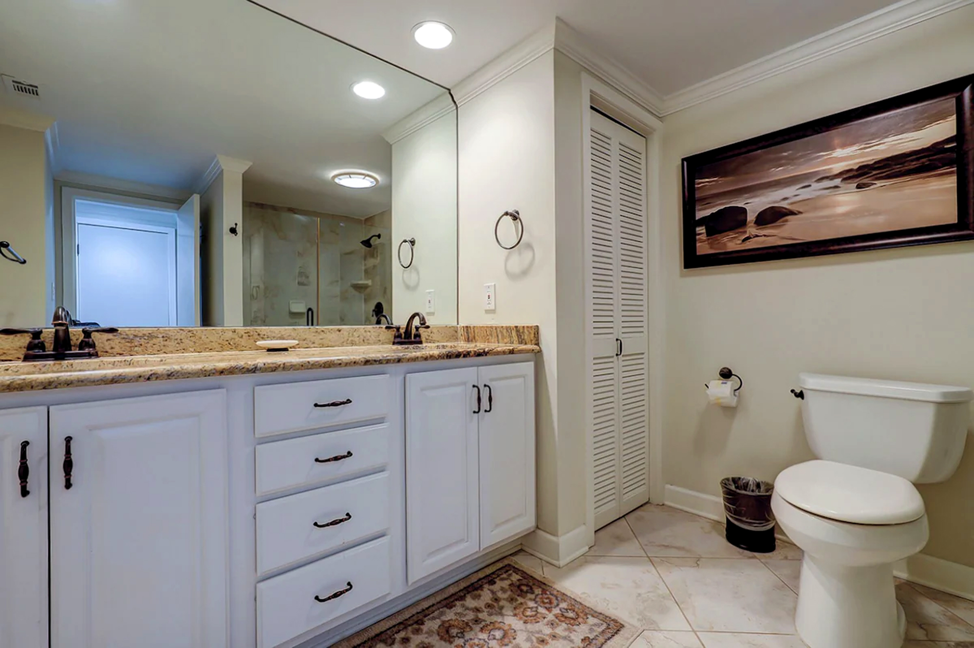 White cabinetry and speckled natural counters create a crisp, clean atmosphere in this spacious bath. Soft overhead glow reflects off the expansive mirror, highlighting the elegant bronze fixtures and earthy tile flooring.