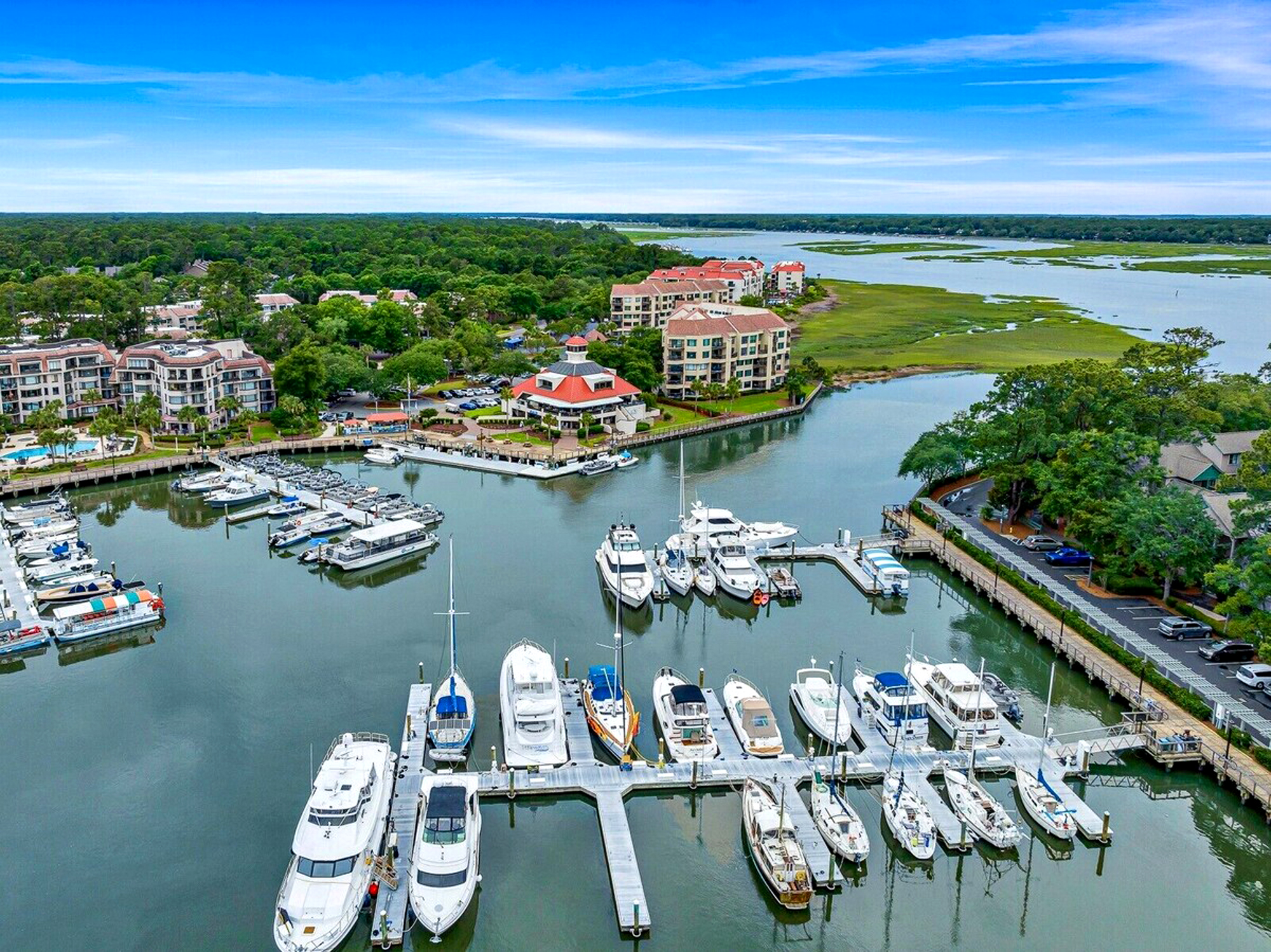 Glistening water surrounds a peaceful marina where sleek boats rest along wooden docks. Verdant trees and coastal architecture frame this serene waterfront scene, perfect for slow mornings watching the tide roll in under a soft blue sky.