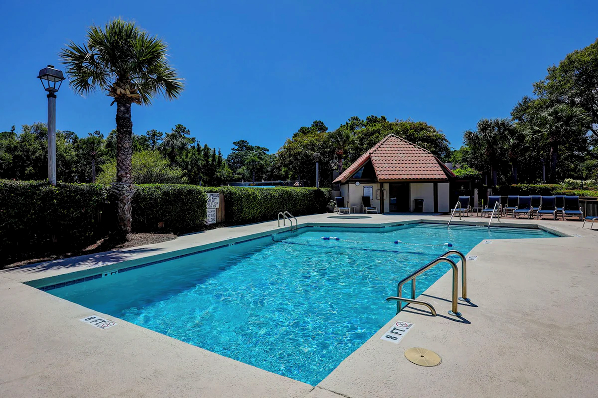 Crystalline ripples dance across the surface of this shimmering swimming pool, bordered by swaying palms and a red tiled cabana. Bright midday rays illuminate the smooth concrete deck, creating a quiet space for a refreshing afternoon dip.