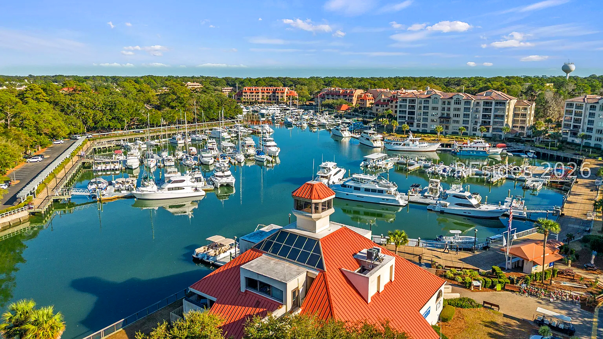Deep blue water fills this vibrant marina, where sleek white yachts rest under a clear afternoon sky. Crimson rooftops and verdant trees frame the docks, creating a peaceful coastal scene that invites you to linger and explore the waterfront.