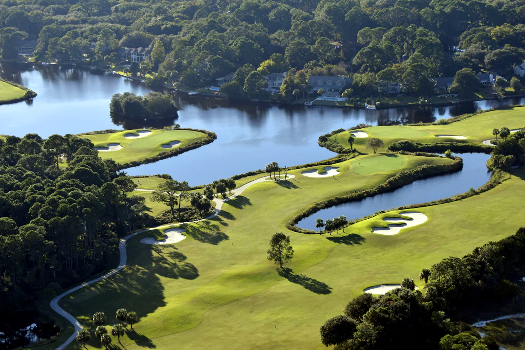 Lush fairways curve gracefully around still, reflective waters and clusters of pines. The late afternoon glow creates soft shadows across the manicured greens, offering a serene landscape for your peaceful getaway.