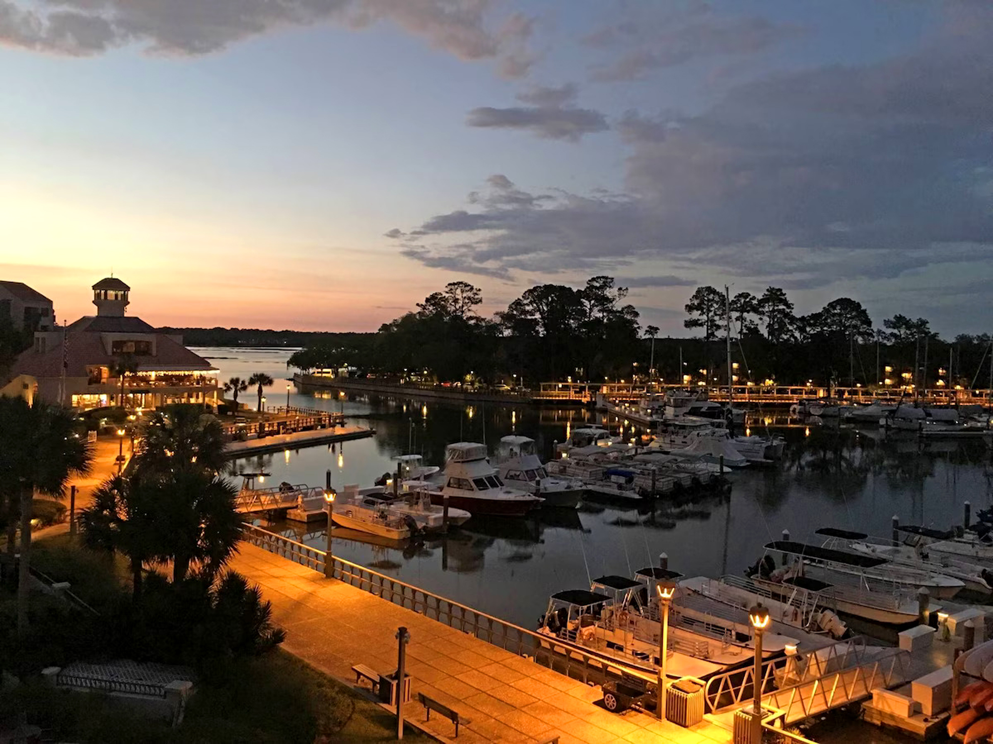 Dusk settles over the quiet marina, where amber streetlamps cast a cozy glow onto the winding boardwalk. Still waters mirror the soft lilac sky and silhouettes of resting boats, offering a peaceful scene for your evening stroll.