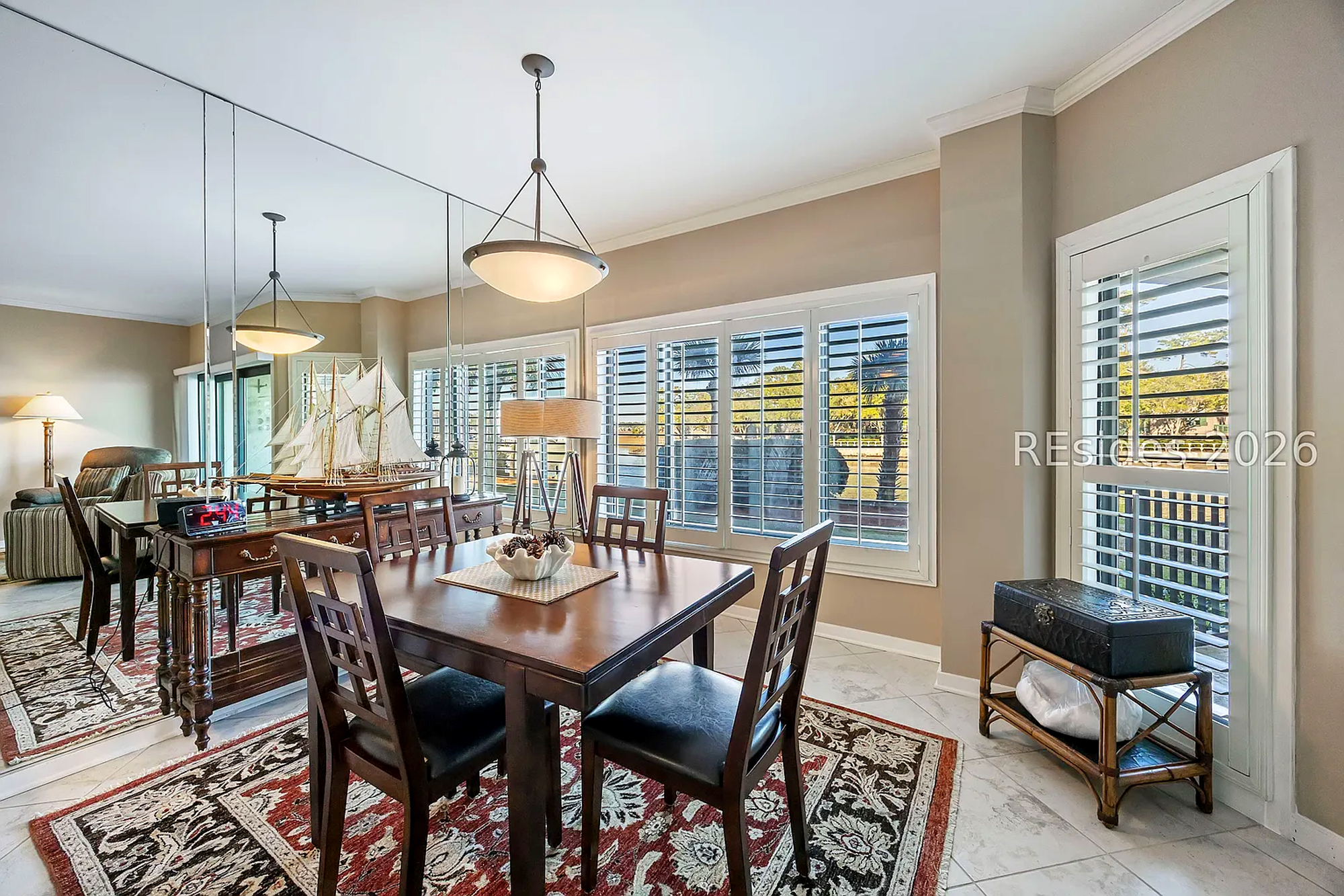 Dark wood grains and nautical accents create a timeless setting for shared meals. Soft afternoon rays filter through plantation shutters, reflecting off the mirrored wall to brighten this elegant dining nook.