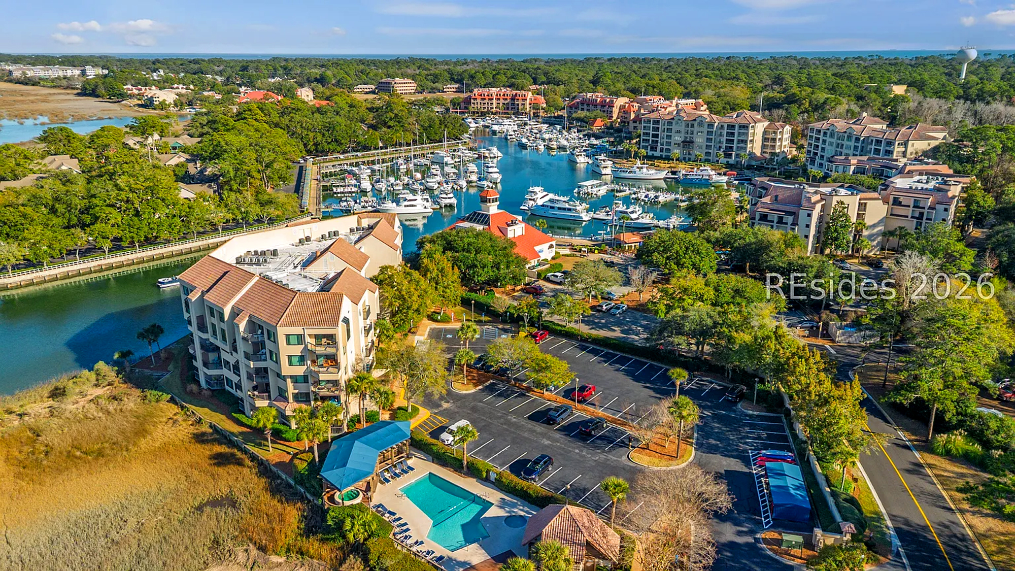 Yacht-filled docks and red-tiled roofs define this coastal scene, where midday brightness illuminates the pool and surrounding greens. Serene water meets the forest edge, creating a peaceful atmosphere for your next stay by the marina.