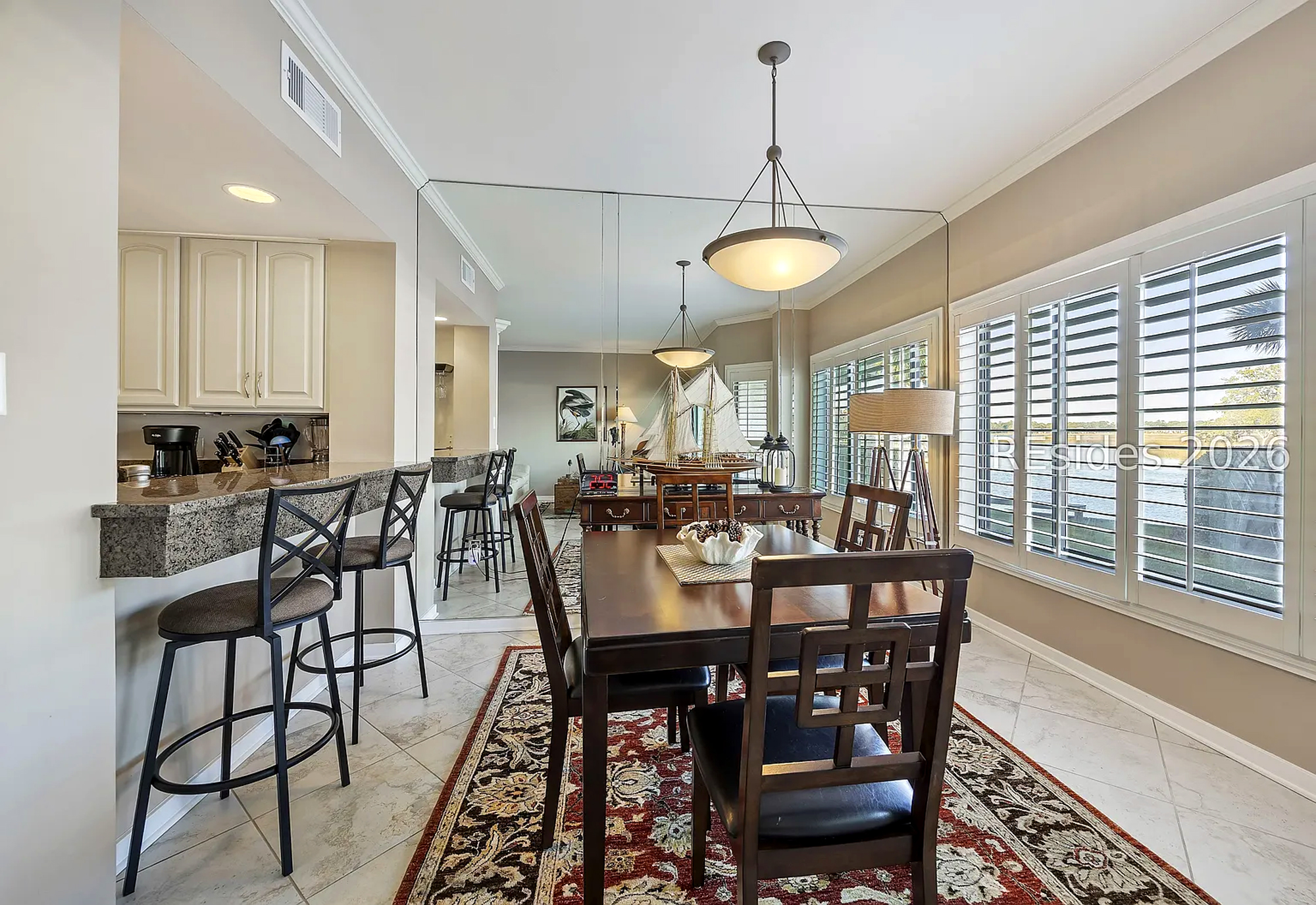 Morning rays filter through plantation shutters, illuminating the dark wood table and intricate oriental rug. This open dining space combines traditional charm with a sleek breakfast bar and mirrored walls to create an airy, social atmosphere.
