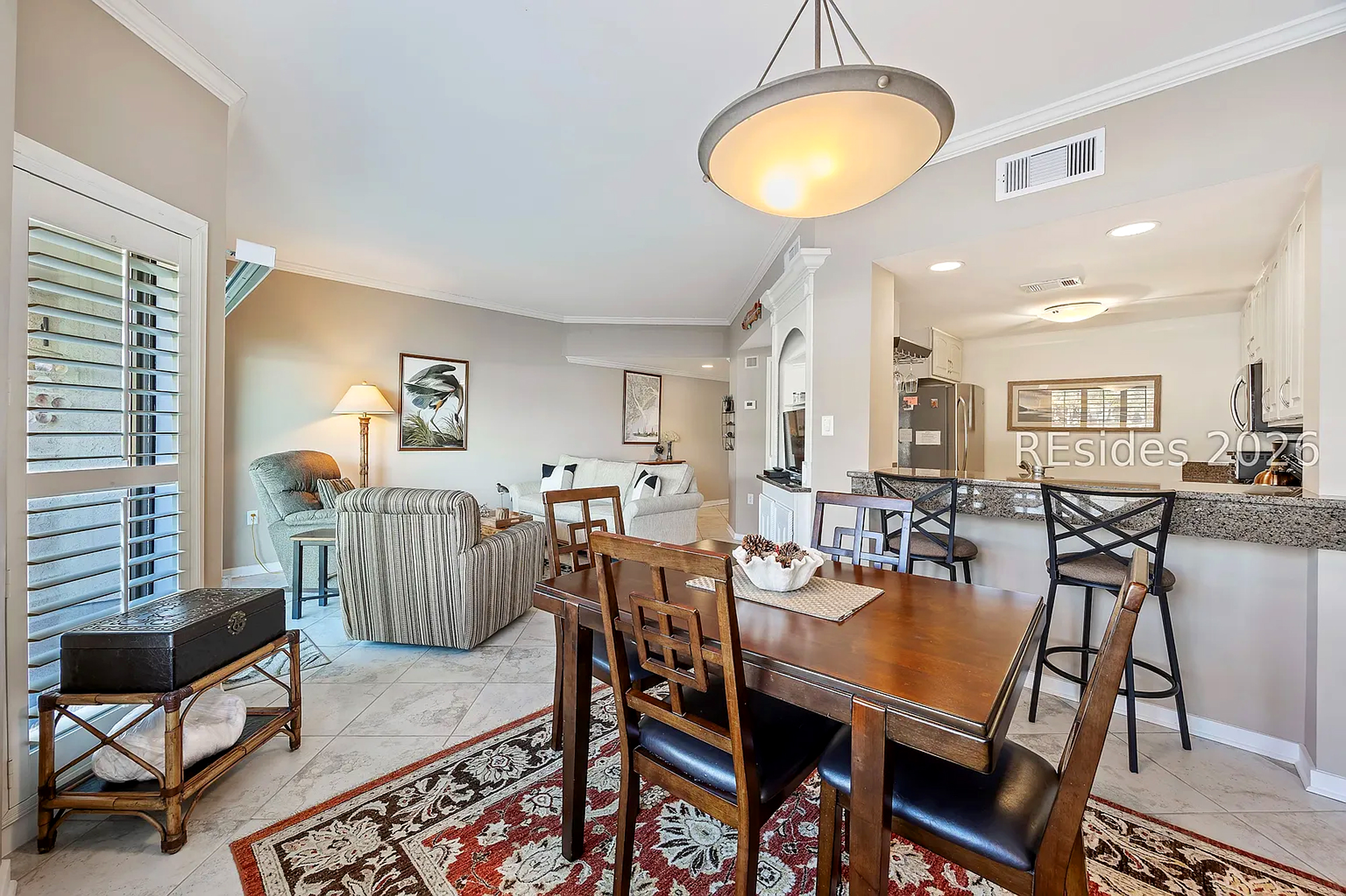 Details highlight wood grains and an ornate rug anchor this airy dining space, where soft overhead glows meet shadows from the louvers. The open layout transitions into a cozy lounge, creating an inviting flow for shared meals and afternoon quiet.