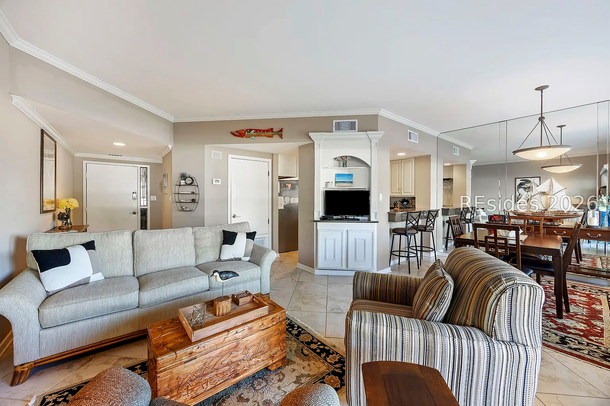Ornate wood carvings and soft, patterned textiles anchor this open living area, where mirrored walls amplify the airy feel. Every corner flows together, from the cozy seating to the nearby kitchen bar, creating a cheerful spot for gathering.