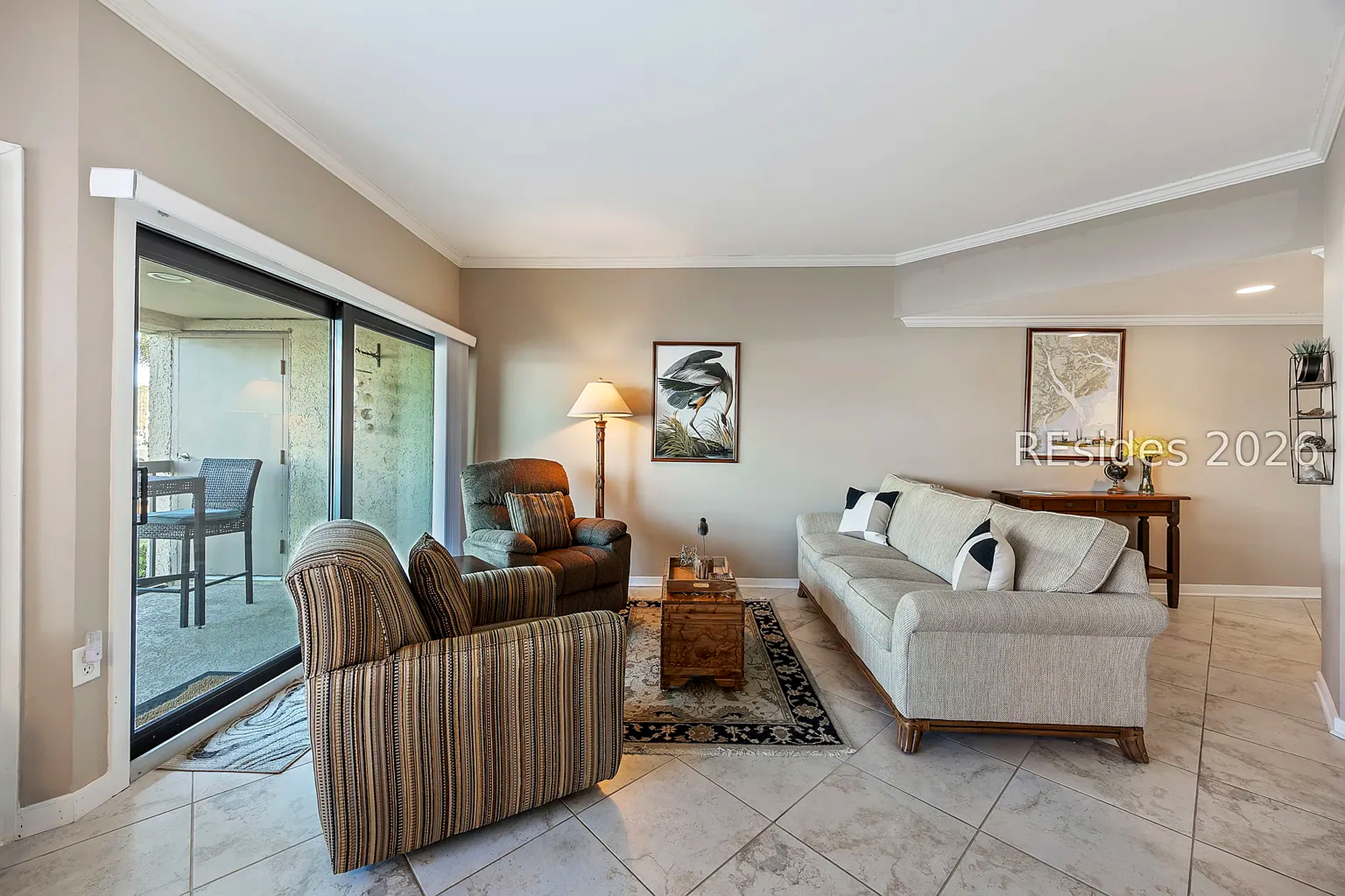 Soft upholstery and striped armchairs create a cozy conversation nook anchored by an intricate rug. Large glass doors pull in the afternoon glow, highlighting the tile floors and artistic wall decor for a calm, inviting atmosphere.