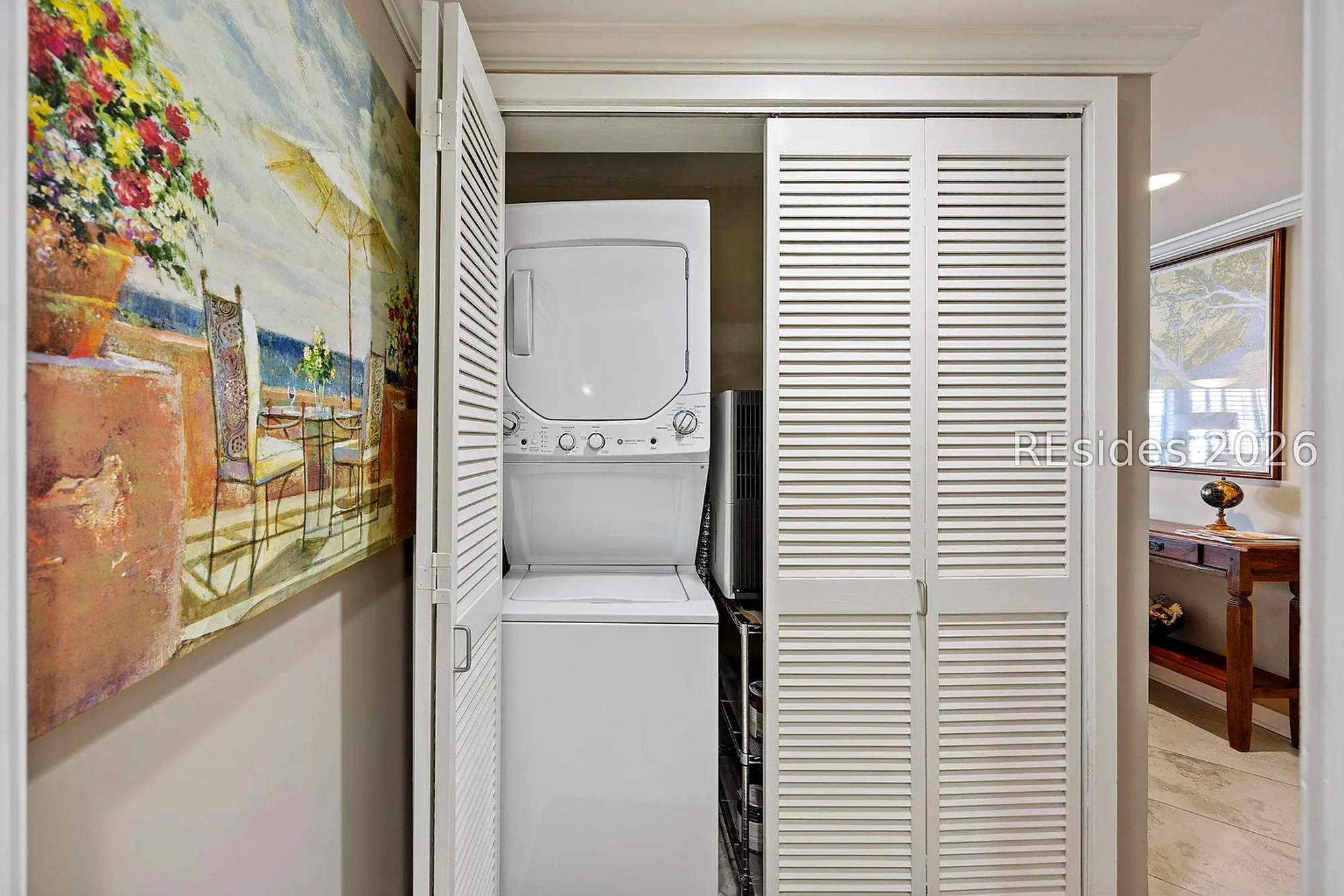 Louvered closet doors swing open to reveal a convenient stacked washer and dryer, combining utility with a clean aesthetic. Bright art and light wood tones create a cheerful atmosphere for your daily routines.