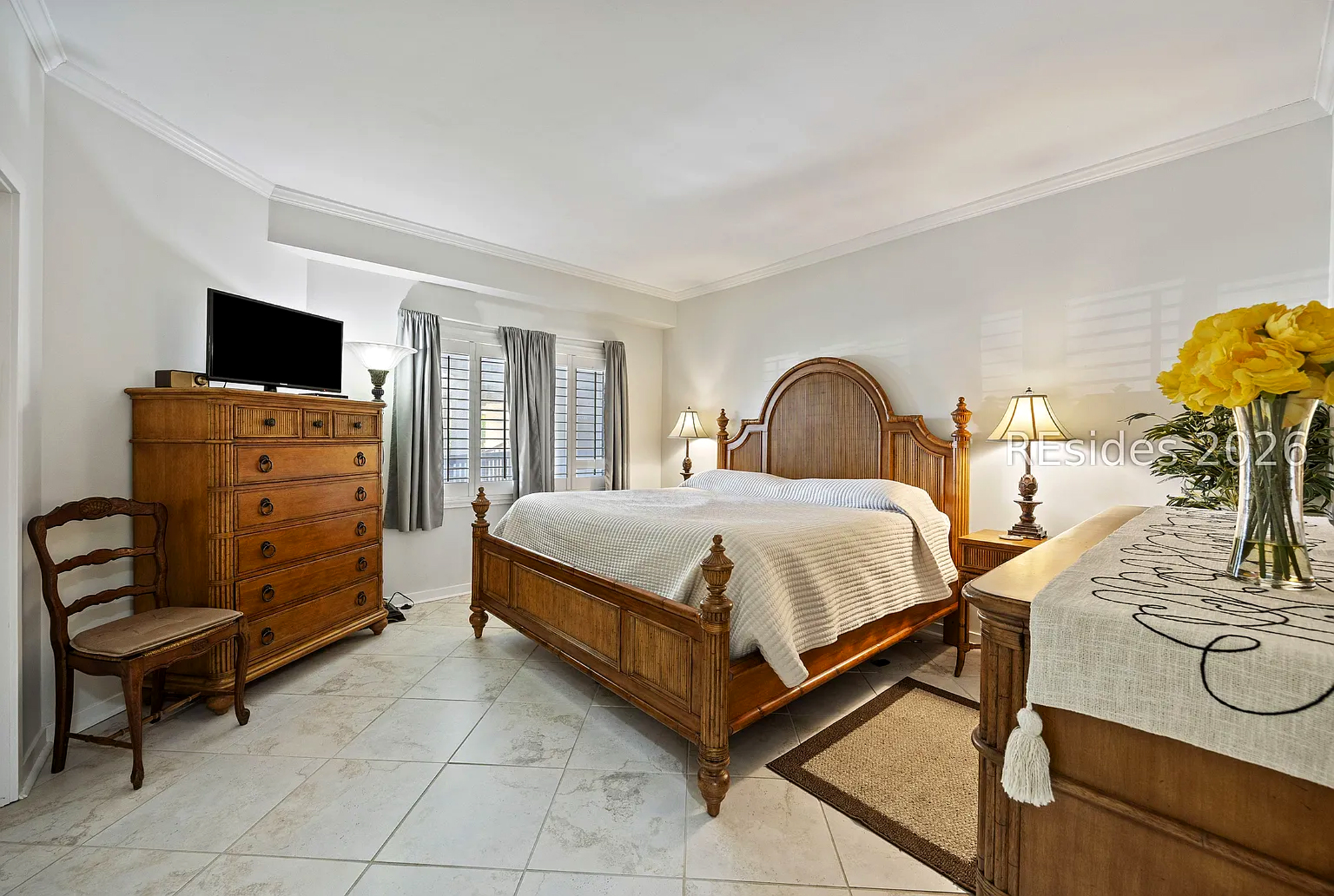 Bamboo-style wood furnishings and a vaulted headboard create a sanctuary of classic comfort. Cool stone tiles balance the room's glow, while fresh yellow blooms add a cheerful pop to this serene, spacious bedroom.