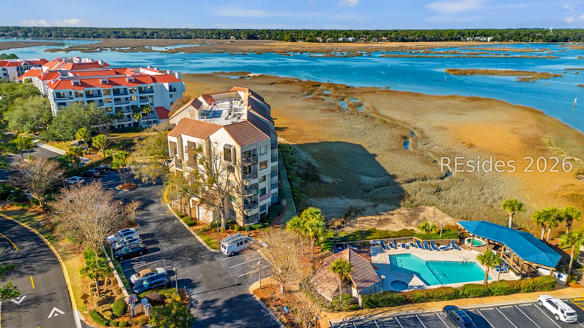 Amber rooflines frame this scenic escape where a pool area meets quiet marshlands and tidal waters. Bright midday light catches the ripples of the cove, offering a peaceful setting for your next coastal getaway.