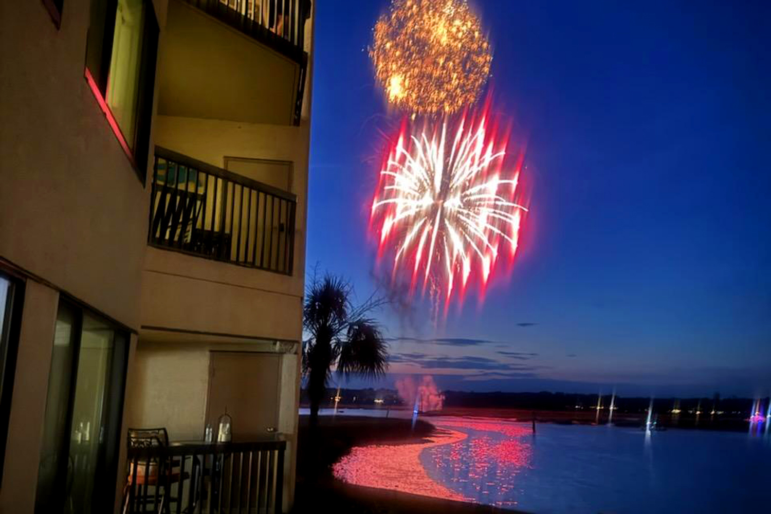 Crimson and gold bursts illuminate the twilight sky, casting a rhythmic glow over the quiet water and your private balcony. This front row view transforms the evening into a spectacular celebration right outside your door.